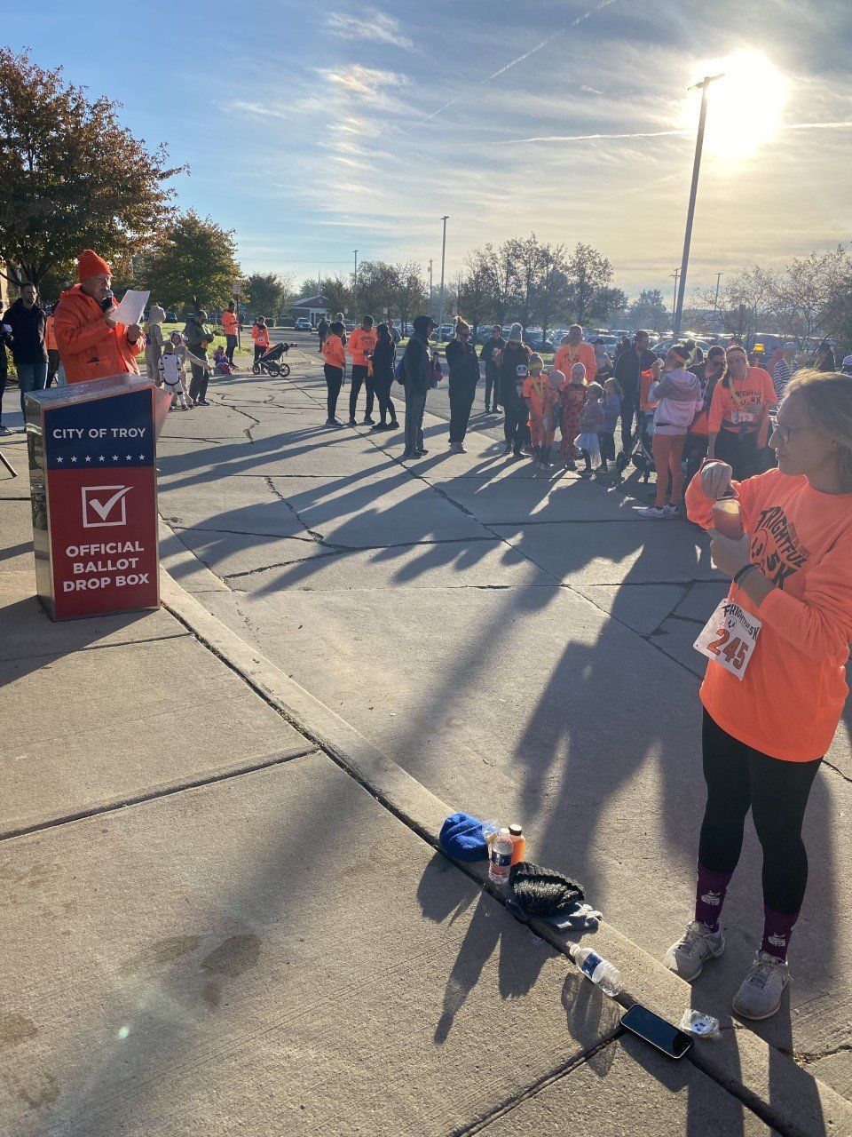 People in orange shirts at a starting line of an outdoor event. A person speaks from a podium. Sun overhead.