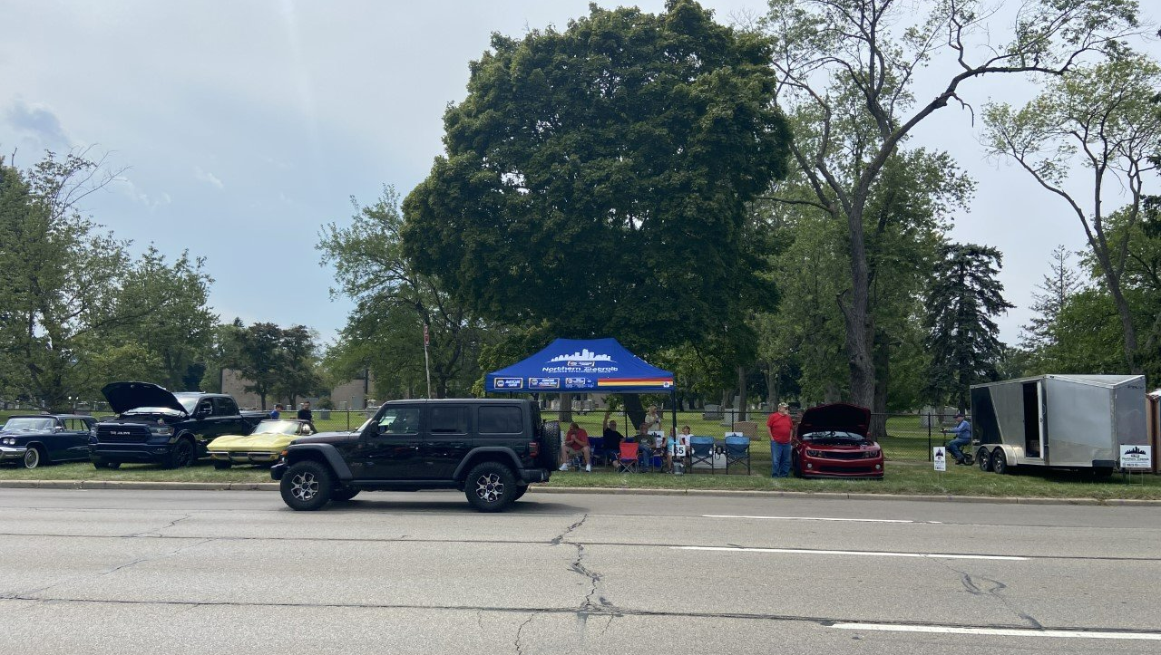 Car show with vehicles parked on the side of a road, people gathered under a tent and near vehicles.