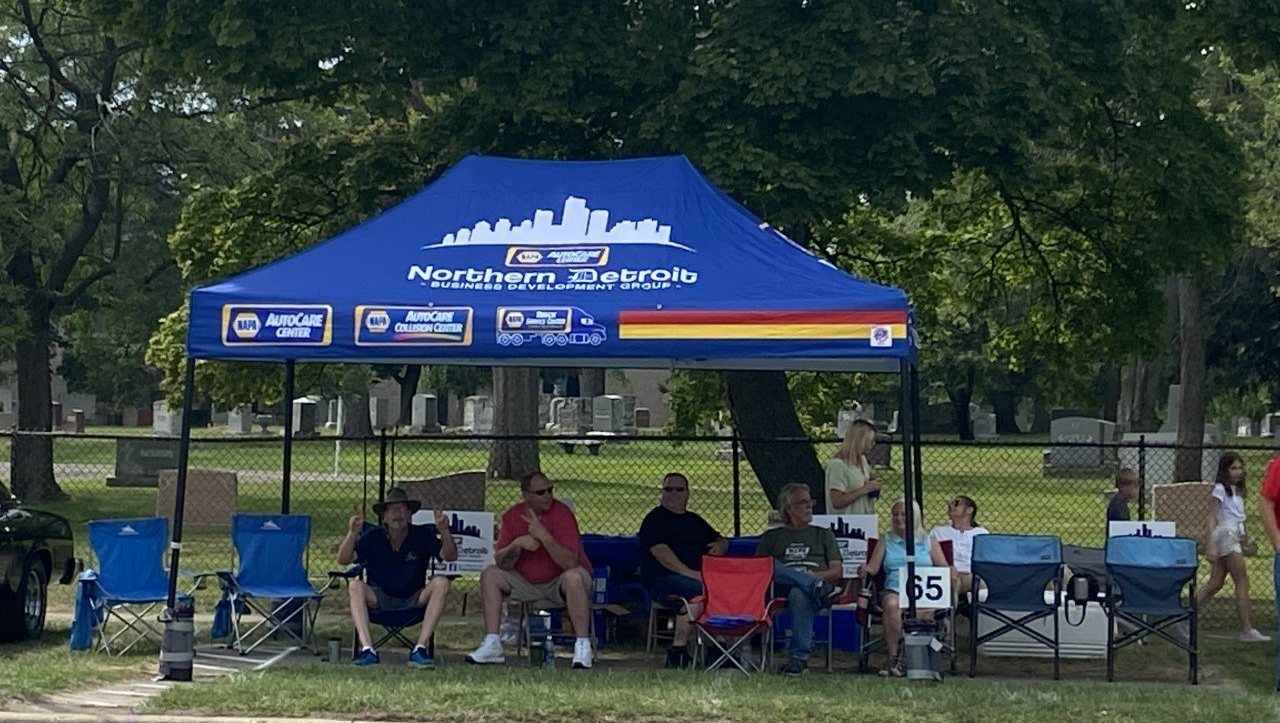 People under a blue canopy with a city skyline logo, seated in folding chairs near a cemetery.