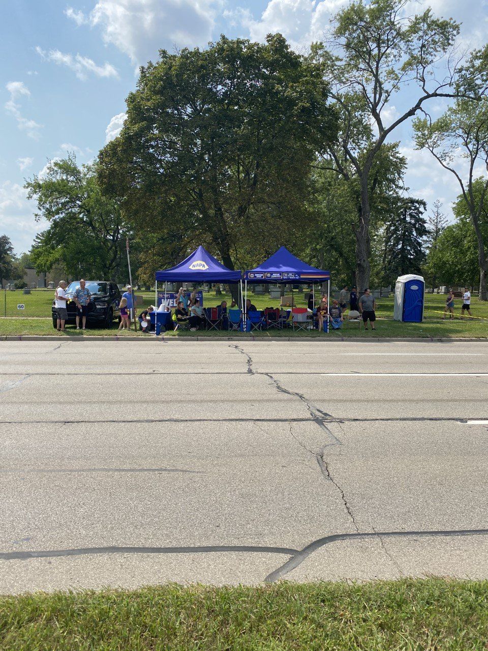Two tents set up roadside with people, likely an event or race support. Blue tents, sunny day.