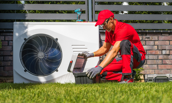 Technician in red uniform inspecting outdoor HVAC unit next to a brick house.