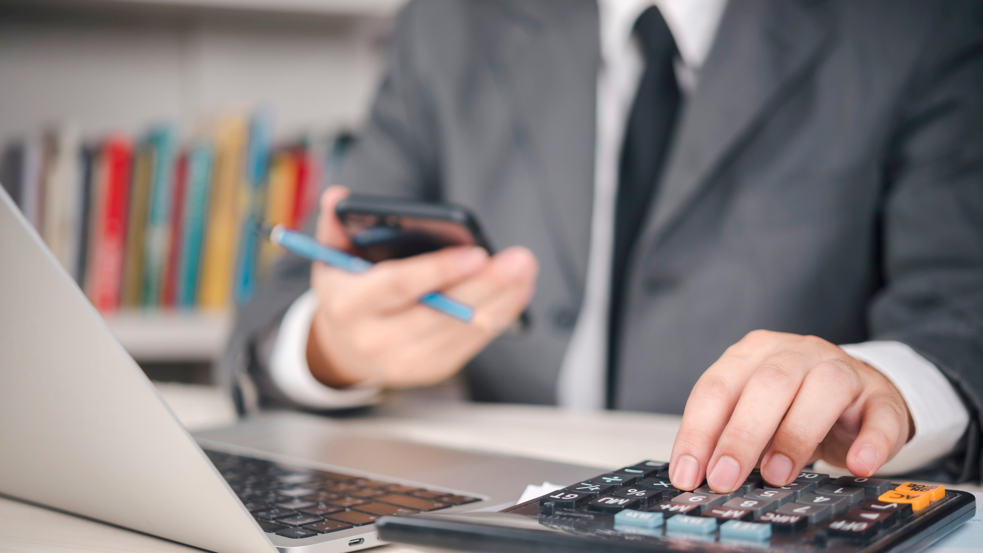 Person in suit using a calculator, holding a phone and pen near a laptop in an office.