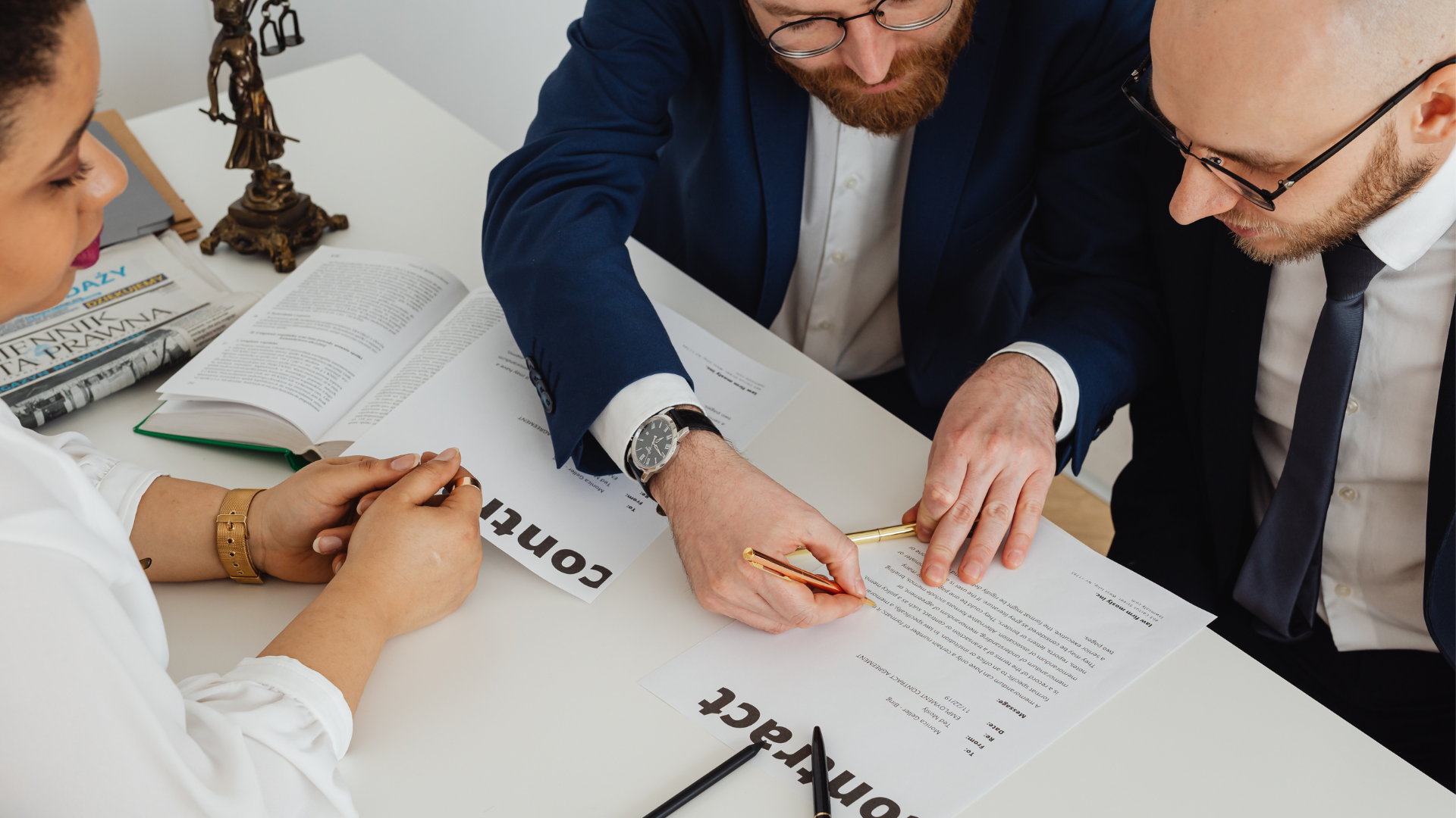 Three people review documents at a table. A man in a suit points a pen at the paperwork.