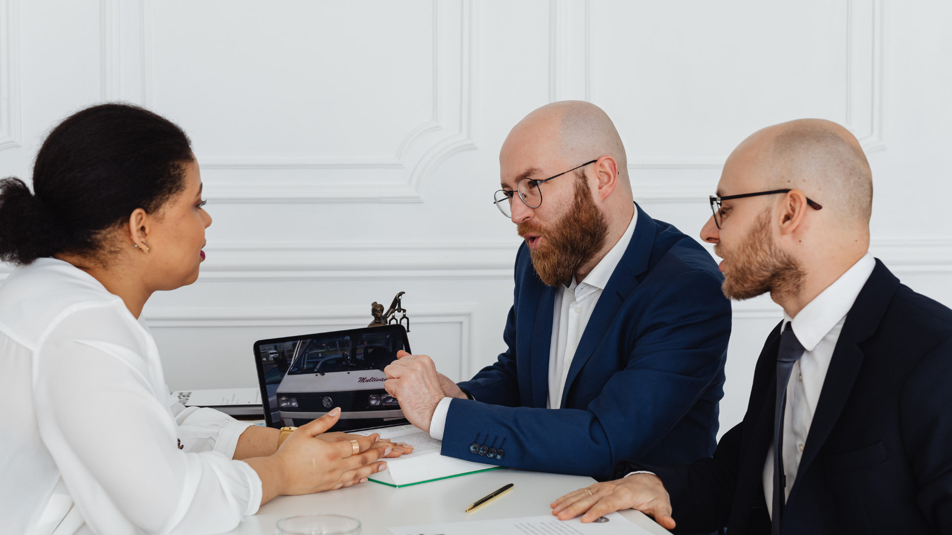 Three people in business attire at a table discussing documents and a laptop, in a bright office.