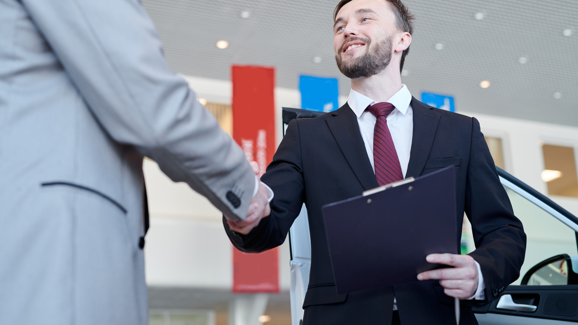 Man in suit shakes hands with another man, car dealership setting. Smiling.