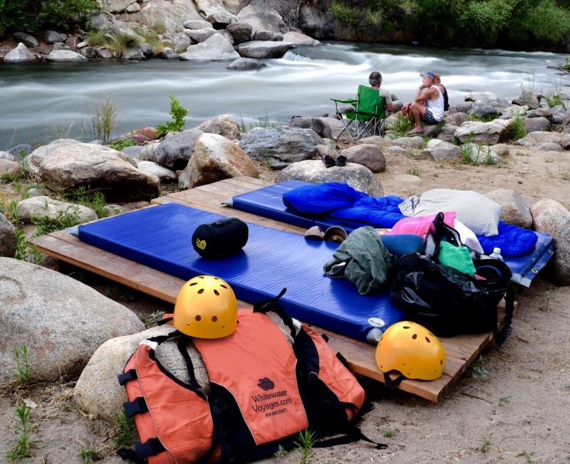 Rafting gear on rocks by a river, with blue pads, orange life jackets, helmets, and people sitting nearby