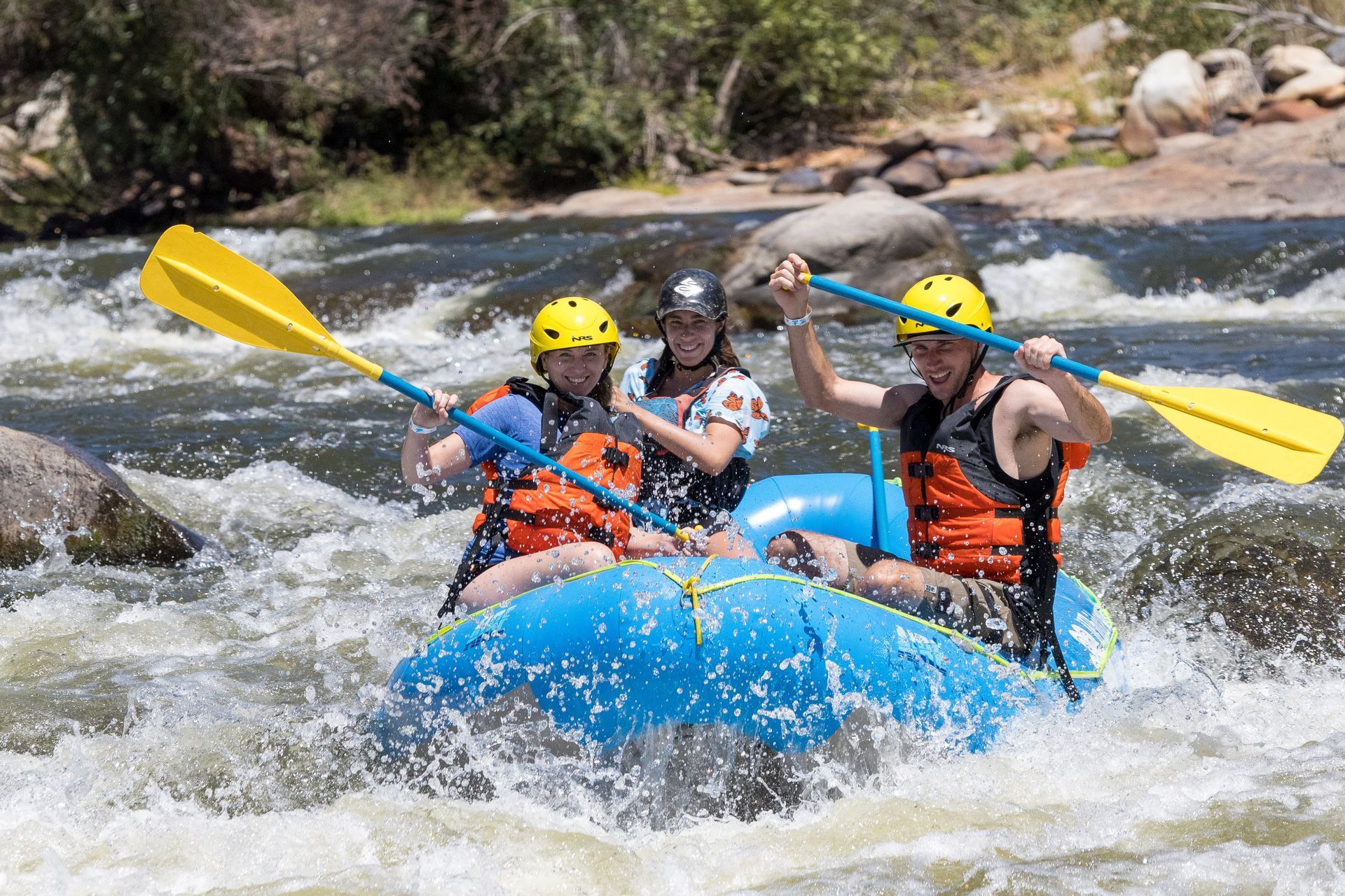 Three rafters in a blue raft paddling through whitewater rapids with yellow paddles