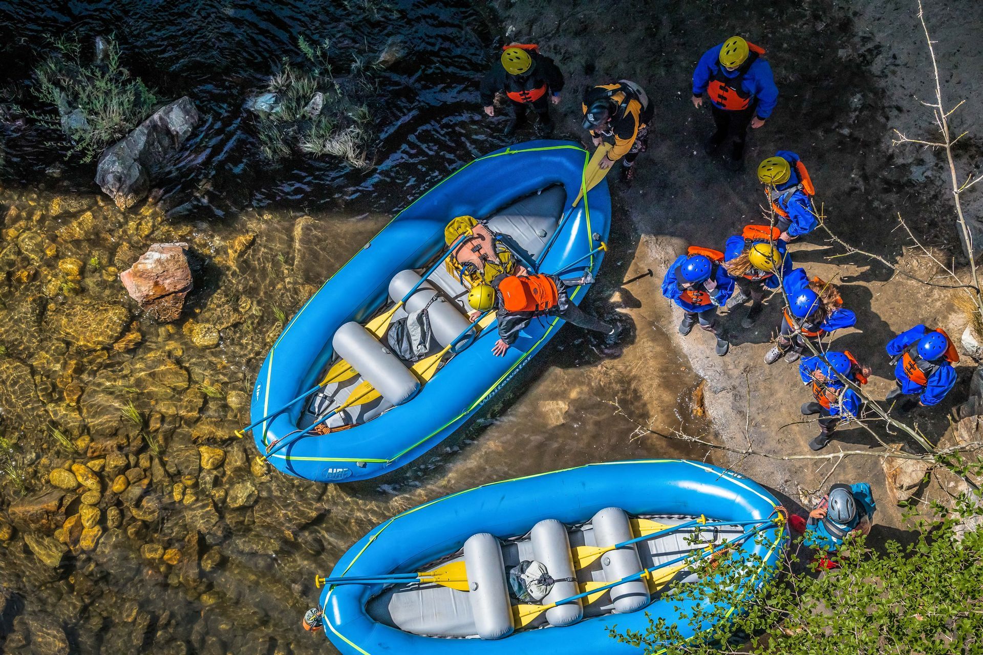 Rafting team in blue rafts on rocky riverbank, with people in orange helmets preparing to launch
