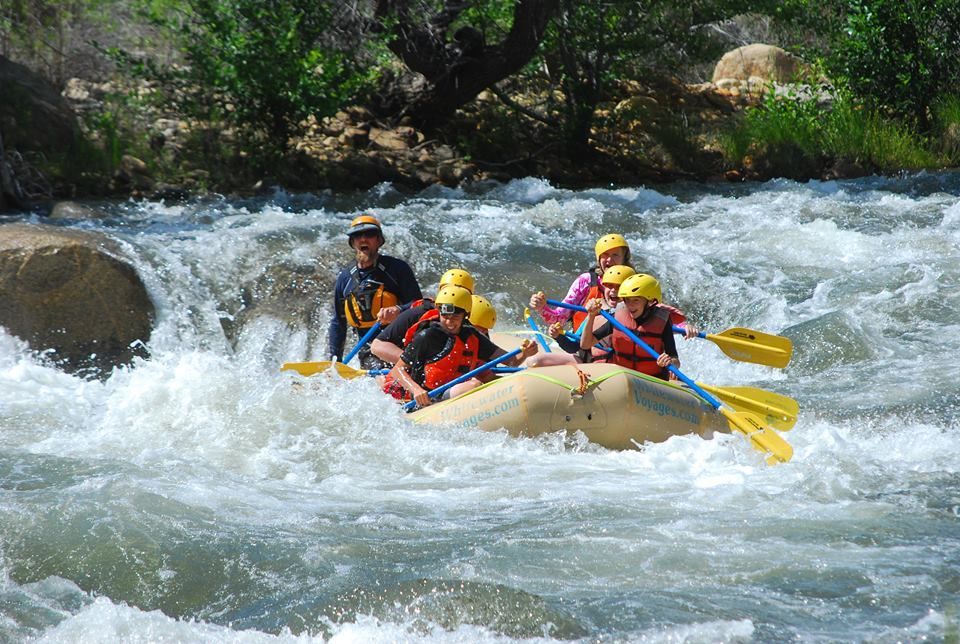 People white water rafting in blue rafts on a river; people wearing helmets and life vests.