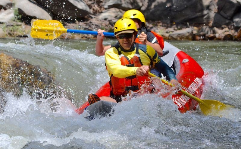 People white water rafting in blue rafts on a river; people wearing helmets and life vests.