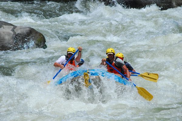 Three rafters in yellow helmets paddle a blue raft through rough whitewater