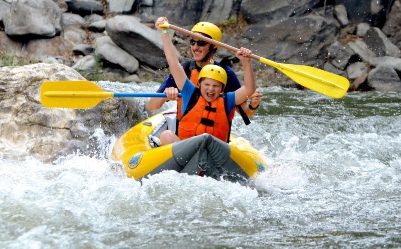 Two people white water rafting, one raising arms in excitement, surrounded by white water.