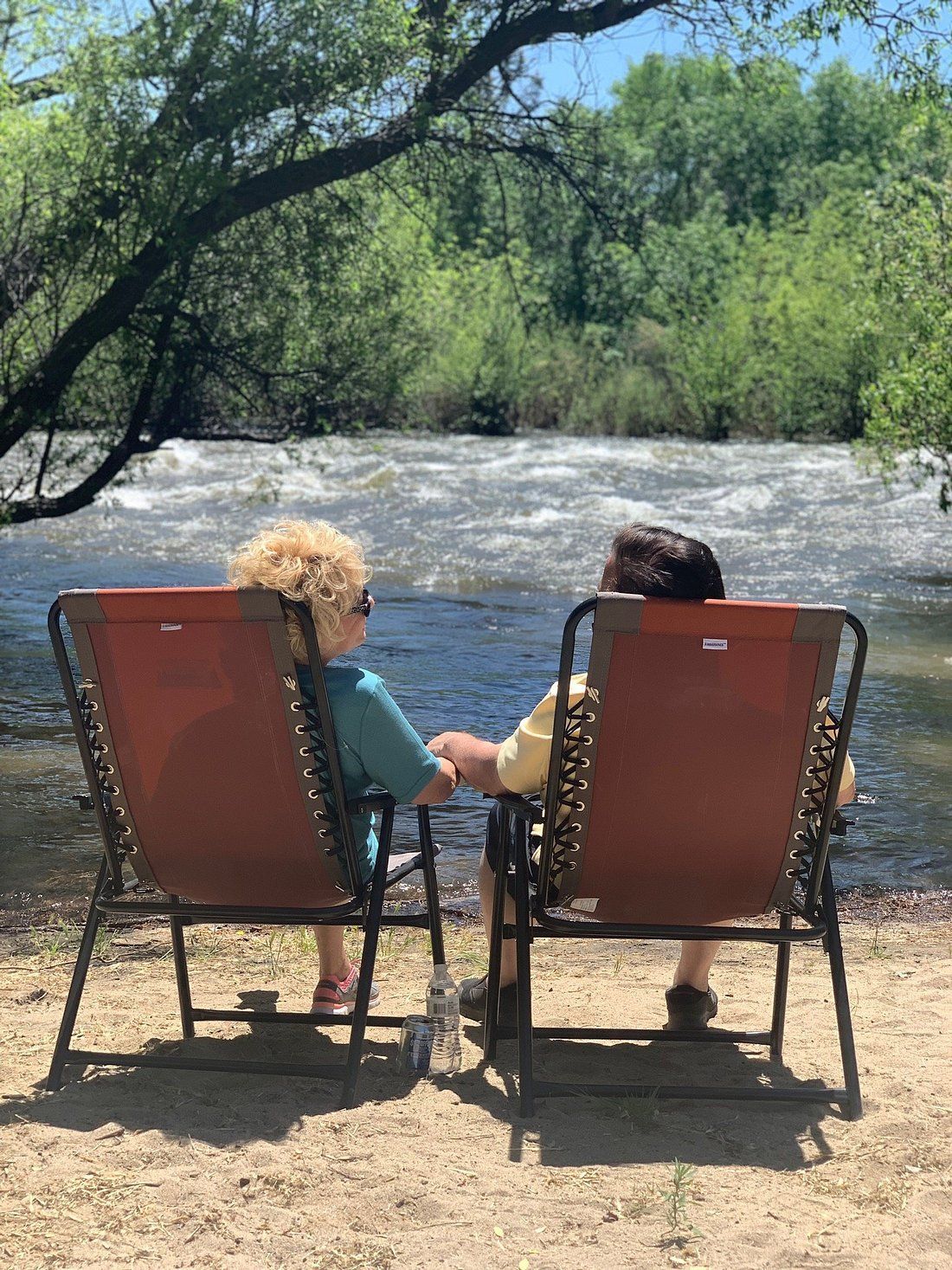Two people are sitting in chairs on the beach looking at a river.