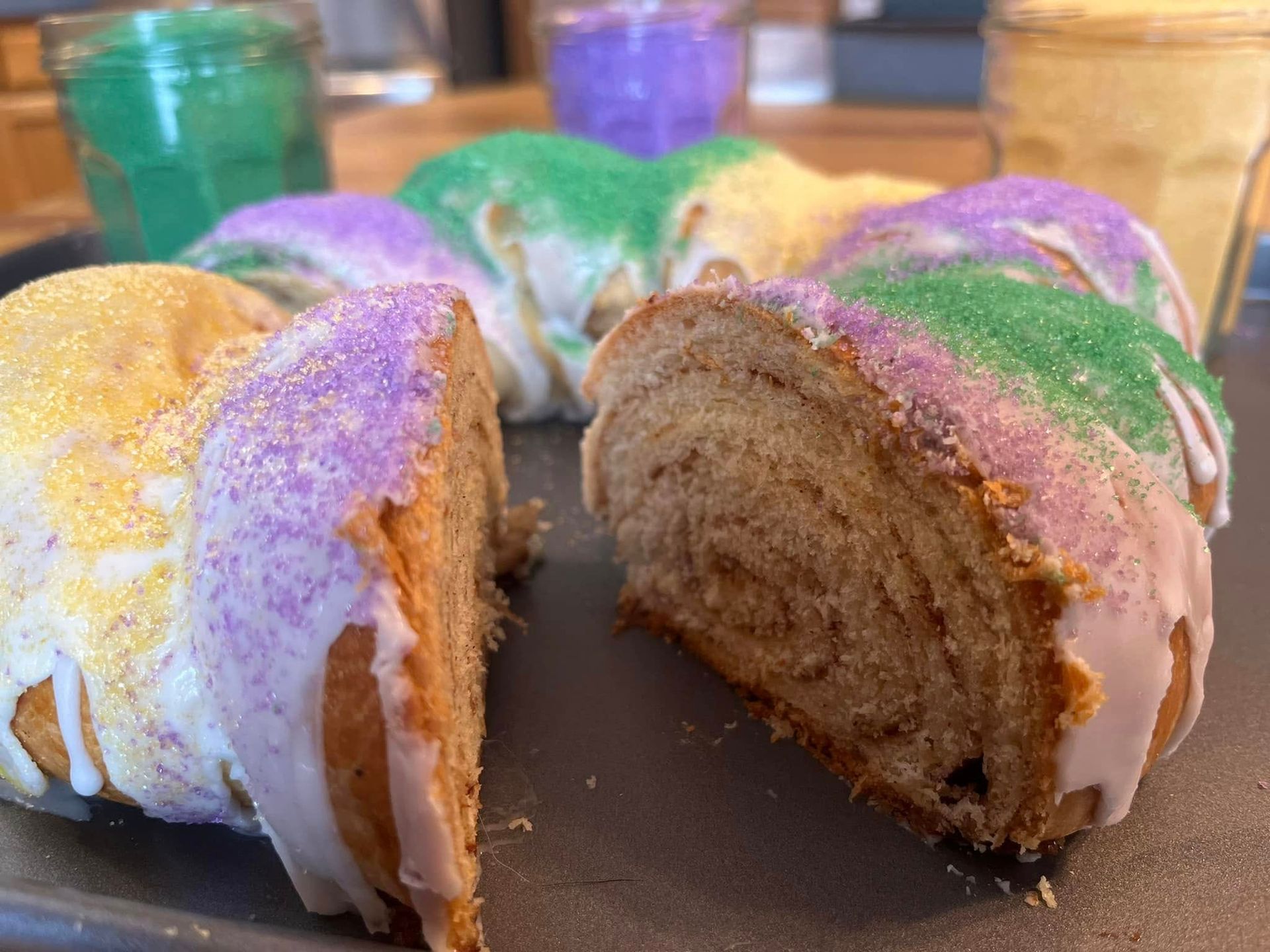 King cake with colored sugar sprinkles and a cut section revealing the cinnamon swirl. Base Camp Bakery Gunnison, Colorado https://share.google/AgqUzUKNzsb0ThlP5, https://www.facebook.com/BaseCampBakery1, https://www.instagram.com/basecampbakery_81230