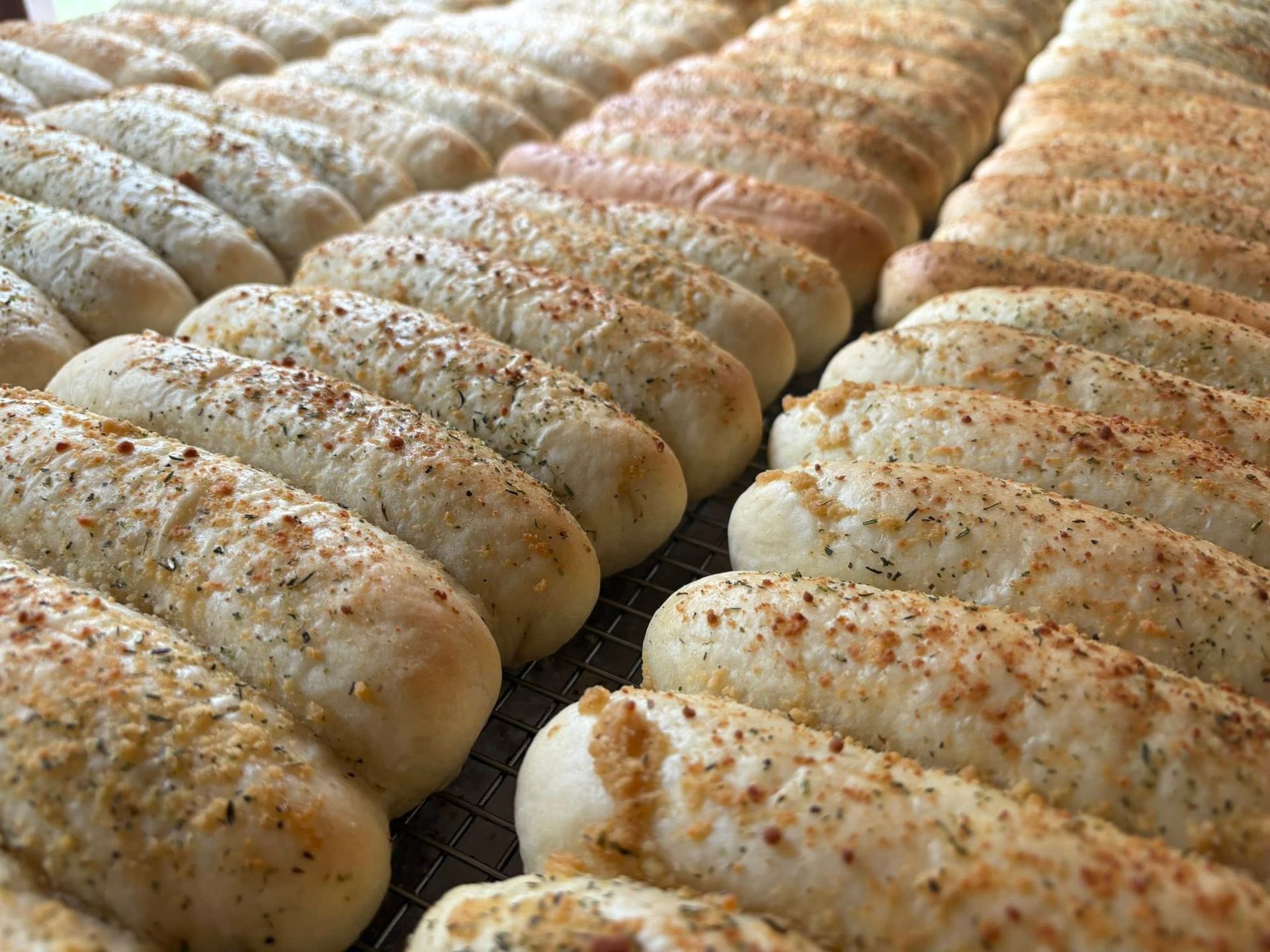 Rows of baked breadsticks, some sprinkled with seasonings, on a cooling rack. Base Camp Bakery Gunnison, Colorado https://share.google/AgqUzUKNzsb0ThlP5, https://www.facebook.com/BaseCampBakery1, https://www.instagram.com/basecampbakery_81230