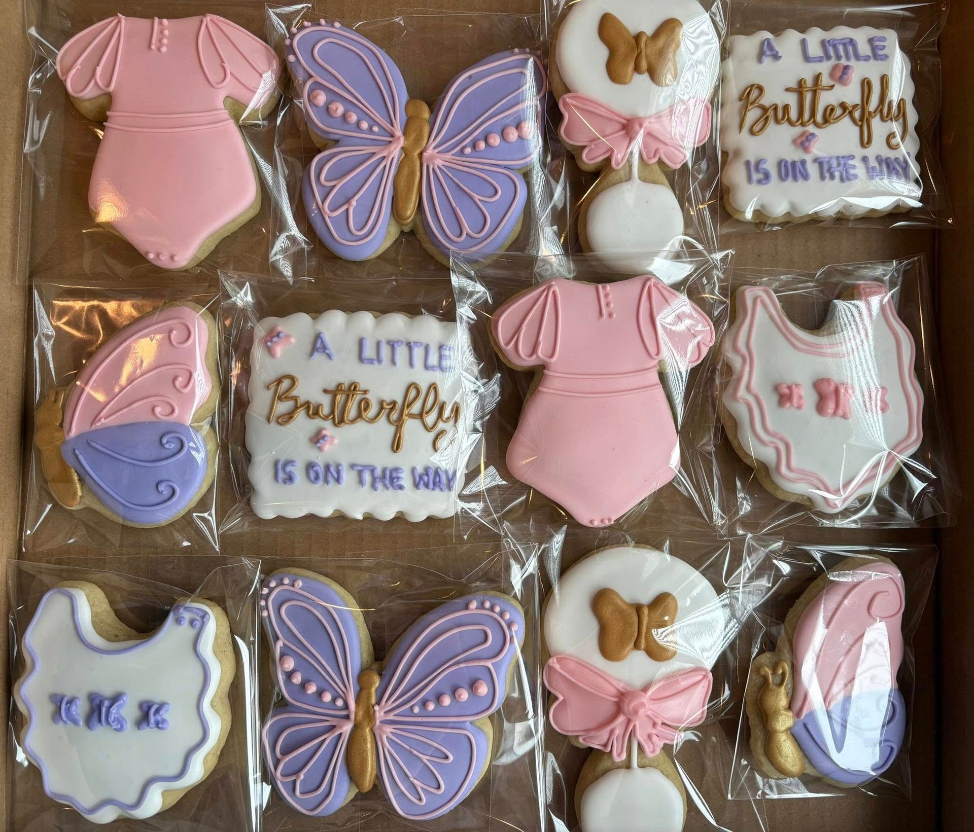 Box of decorated baby shower cookies: butterflies, onesies, bibs, and rattles in pink, purple, and gold. Base Camp Bakery Gunnison, Colorado https://share.google/AgqUzUKNzsb0ThlP5, https://www.facebook.com/BaseCampBakery1, https://www.instagram.com/basecampbakery_81230