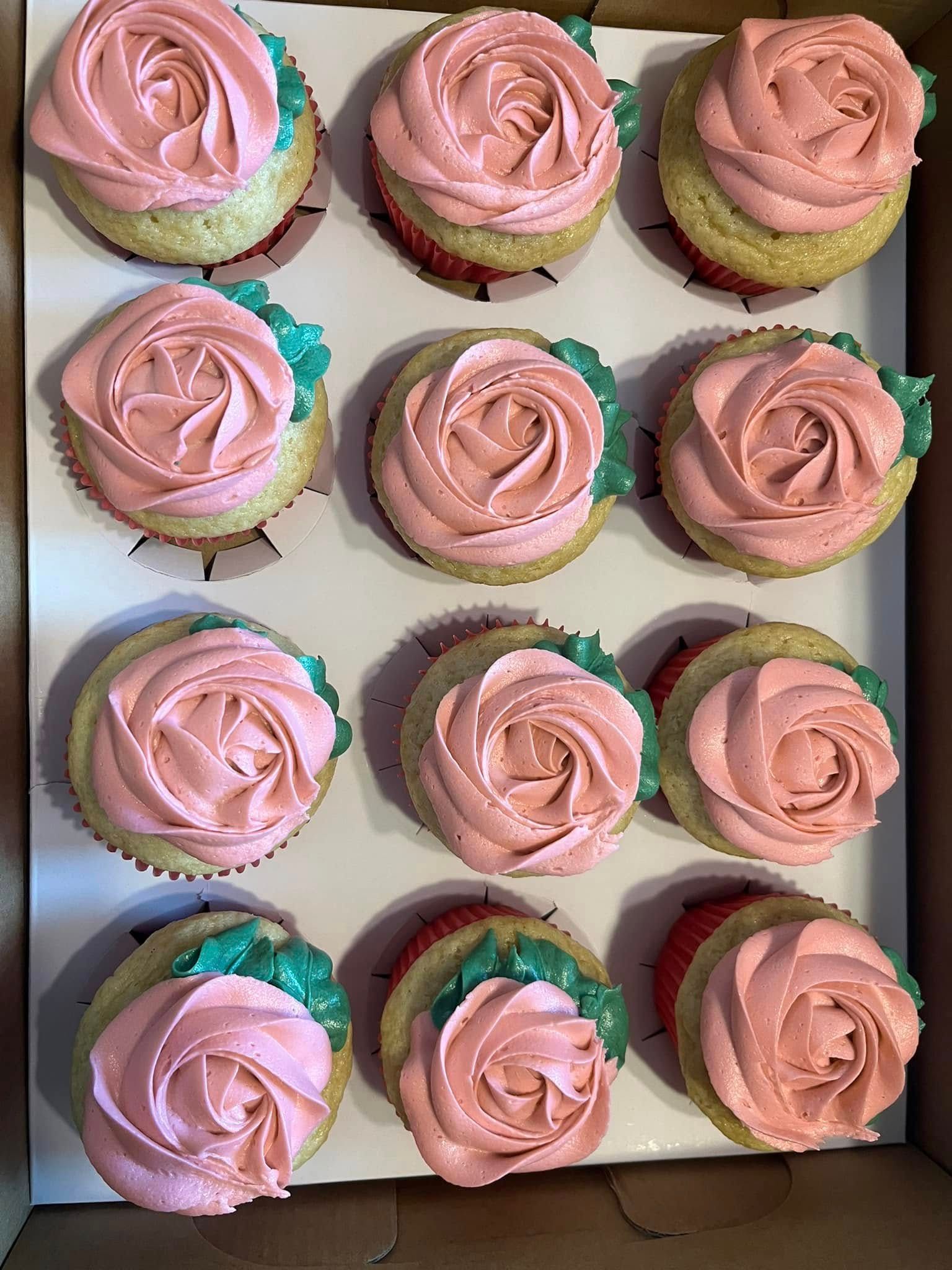 Box of a dozen cupcakes with pink rose frosting and green leaf accents. Base Camp Bakery Gunnison, Colorado https://share.google/AgqUzUKNzsb0ThlP5, https://www.facebook.com/BaseCampBakery1, https://www.instagram.com/basecampbakery_81230