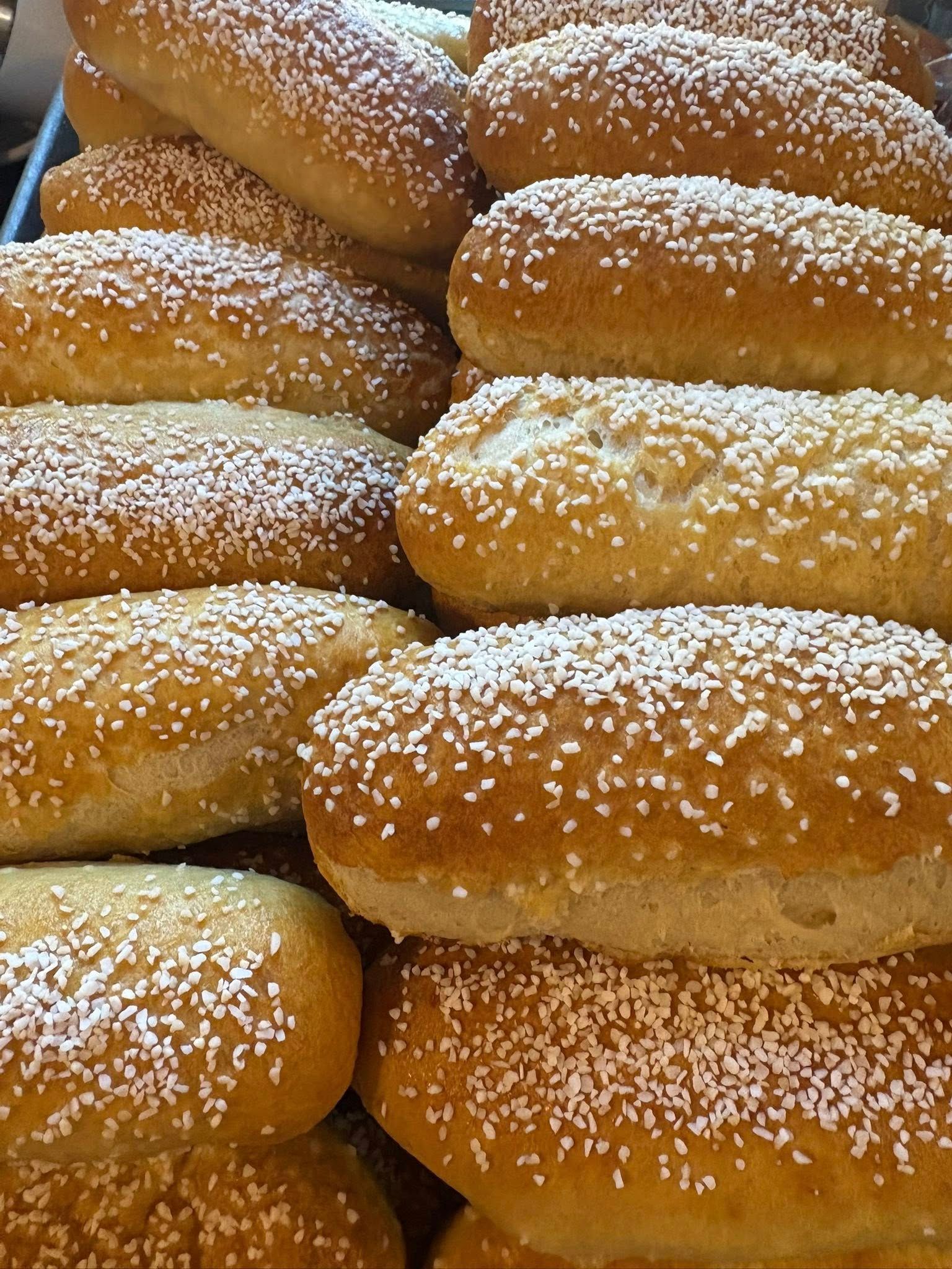 Golden-brown, oblong-shaped bread rolls, covered in white sprinkles, stacked in a display. Base Camp Bakery Gunnison, Colorado https://share.google/AgqUzUKNzsb0ThlP5, https://www.facebook.com/BaseCampBakery1, https://www.instagram.com/basecampbakery_81230