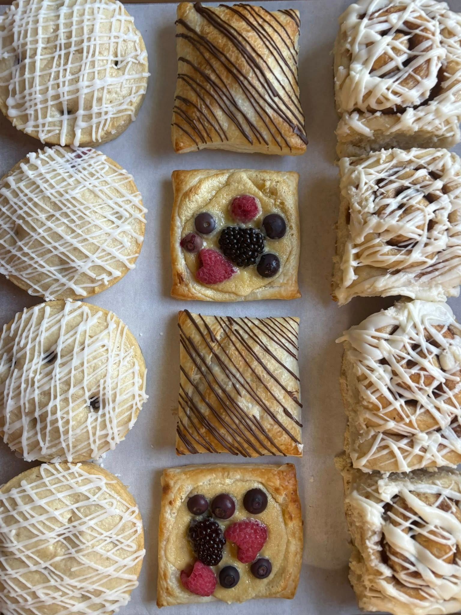 Pastries arranged in rows: muffins drizzled, rectangular danishes, and cinnamon rolls. Base Camp Bakery Gunnison, Colorado https://share.google/AgqUzUKNzsb0ThlP5, https://www.facebook.com/BaseCampBakery1, https://www.instagram.com/basecampbakery_81230