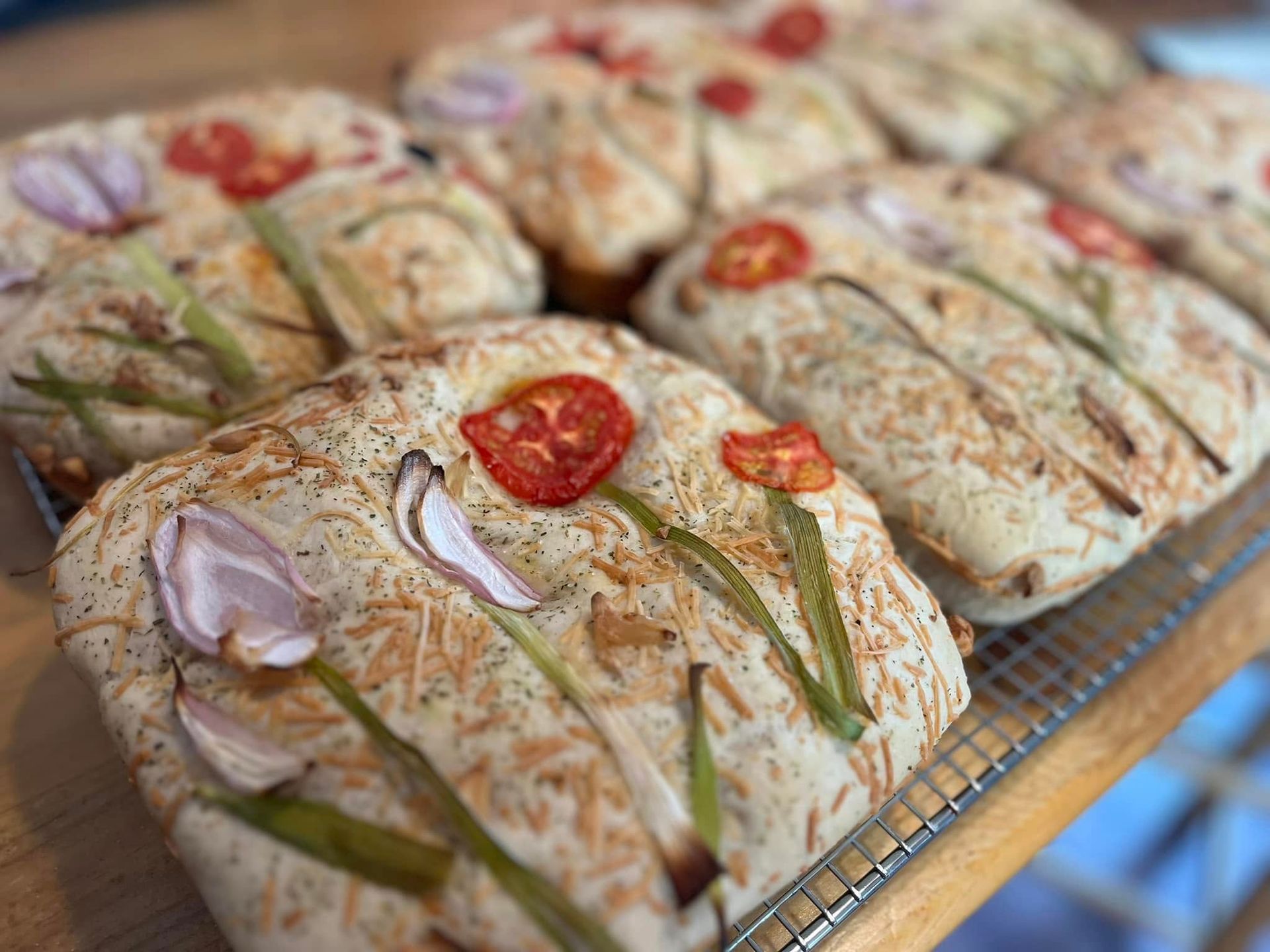 Focaccia bread decorated with tomatoes, onions, and chives, baked on a wire rack. Base Camp Bakery Gunnison, Colorado https://share.google/AgqUzUKNzsb0ThlP5, https://www.facebook.com/BaseCampBakery1, https://www.instagram.com/basecampbakery_81230