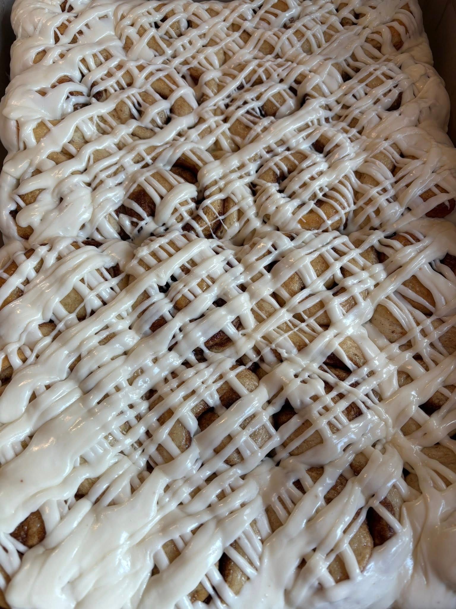 Overhead view of frosted cinnamon rolls in a baking pan, drizzled with white icing. Base Camp Bakery Gunnison, Colorado https://share.google/AgqUzUKNzsb0ThlP5, https://www.facebook.com/BaseCampBakery1, https://www.instagram.com/basecampbakery_81230