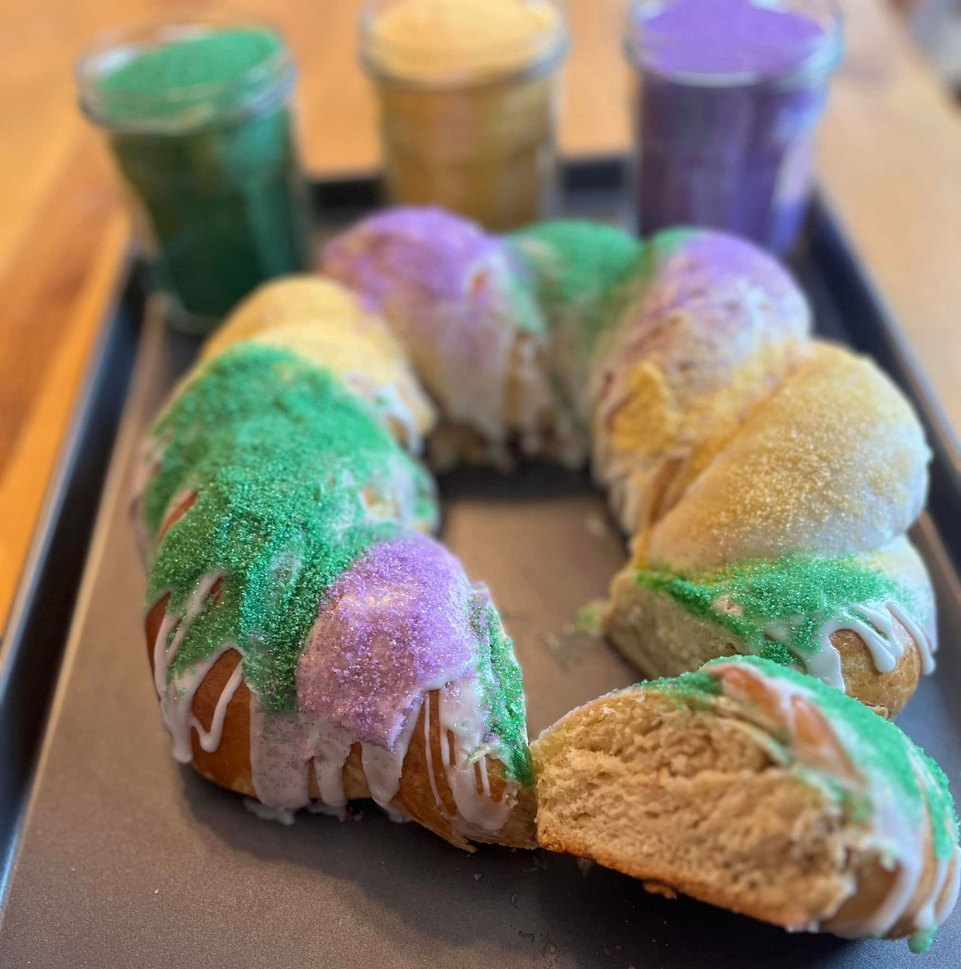 King cake with purple, green, and yellow sugar, served with colored sugar jars on a tray. Base Camp Bakery Gunnison, Colorado https://share.google/AgqUzUKNzsb0ThlP5, https://www.facebook.com/BaseCampBakery1, https://www.instagram.com/basecampbakery_81230
