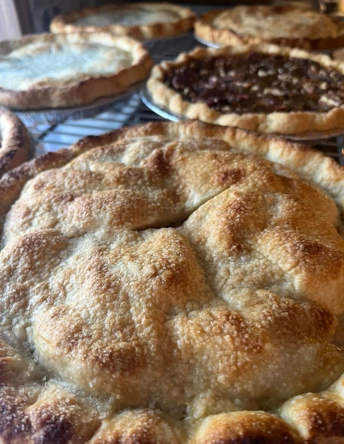 Close-up of a golden-brown pie with several other pies in the background, all on a metal rack. Base Camp Bakery Gunnison, Colorado https://share.google/AgqUzUKNzsb0ThlP5, https://www.facebook.com/BaseCampBakery1, https://www.instagram.com/basecampbakery_81230