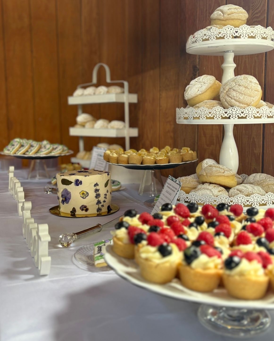Dessert table: tiered stands with cookies, tarts with berries, cake, and mini pastries on a white tablecloth. Base Camp Bakery Gunnison, Colorado https://share.google/AgqUzUKNzsb0ThlP5, https://www.facebook.com/BaseCampBakery1, https://www.instagram.com/basecampbakery_81230