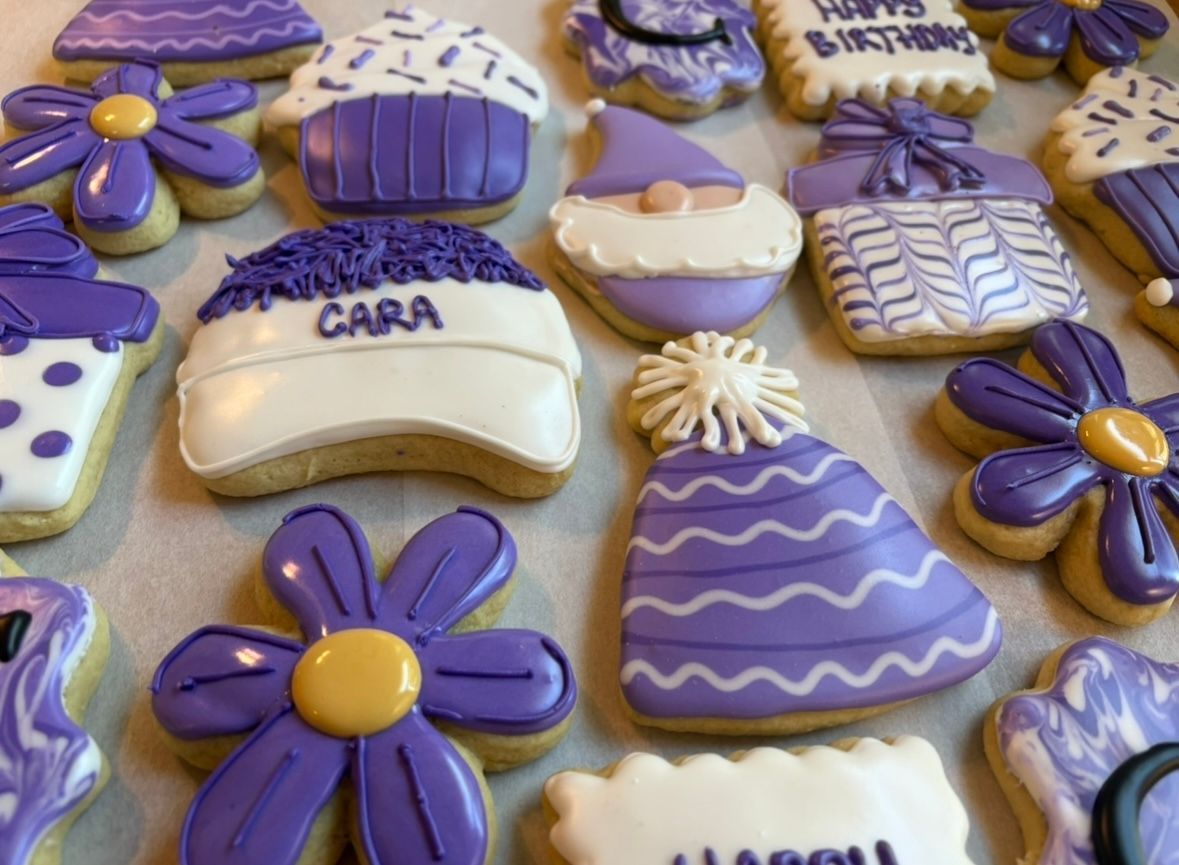 Purple frosted cookies: cupcakes, flowers, hats, a gnome, and a gift box, decorated for a celebration. Base Camp Bakery Gunnison, Colorado https://share.google/AgqUzUKNzsb0ThlP5, https://www.facebook.com/BaseCampBakery1, https://www.instagram.com/basecampbakery_81230