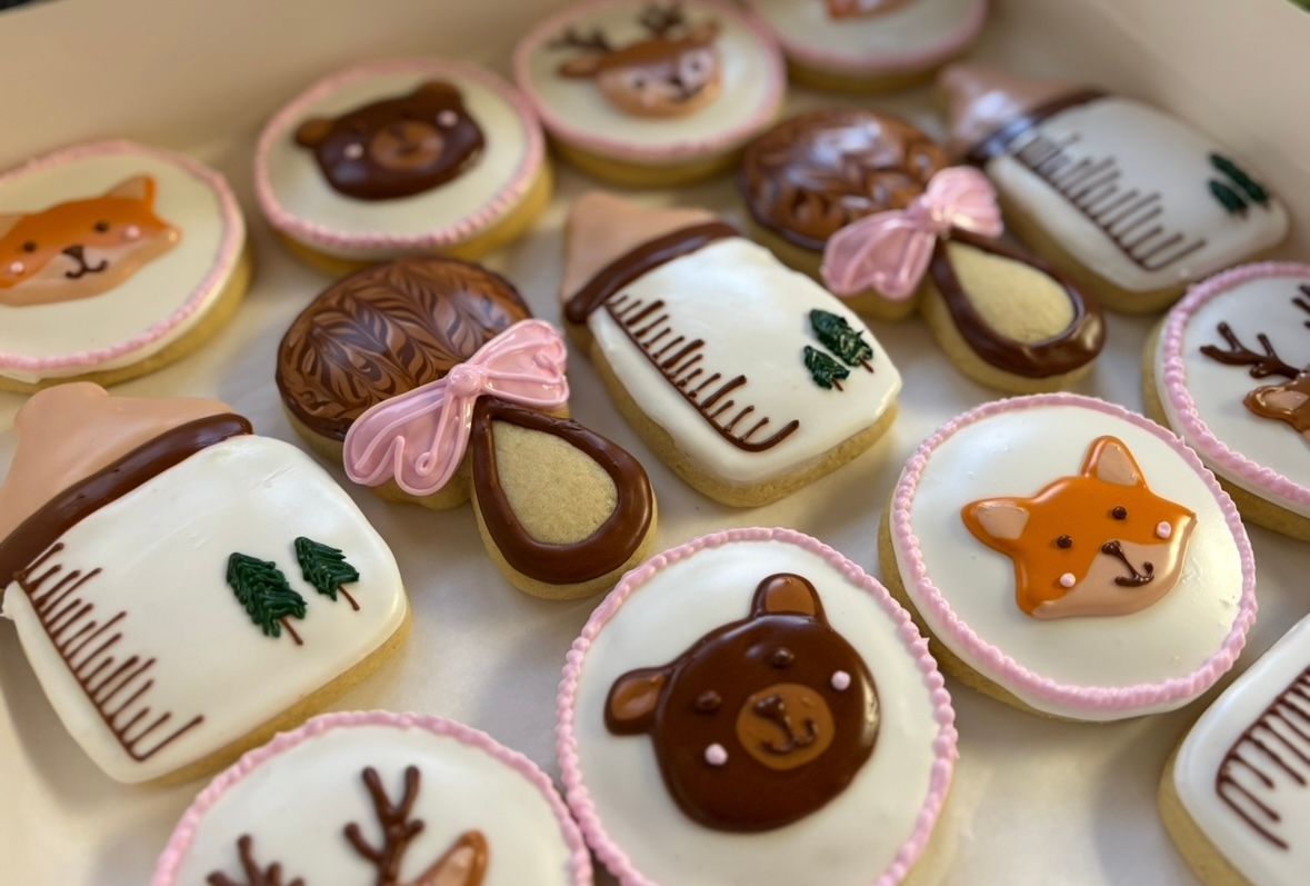 Box of decorated baby-themed cookies: bottles, rattles, fox, bear, and deer faces, white and pink frosting. Base Camp Bakery Gunnison, Colorado https://share.google/AgqUzUKNzsb0ThlP5, https://www.facebook.com/BaseCampBakery1, https://www.instagram.com/basecampbakery_81230