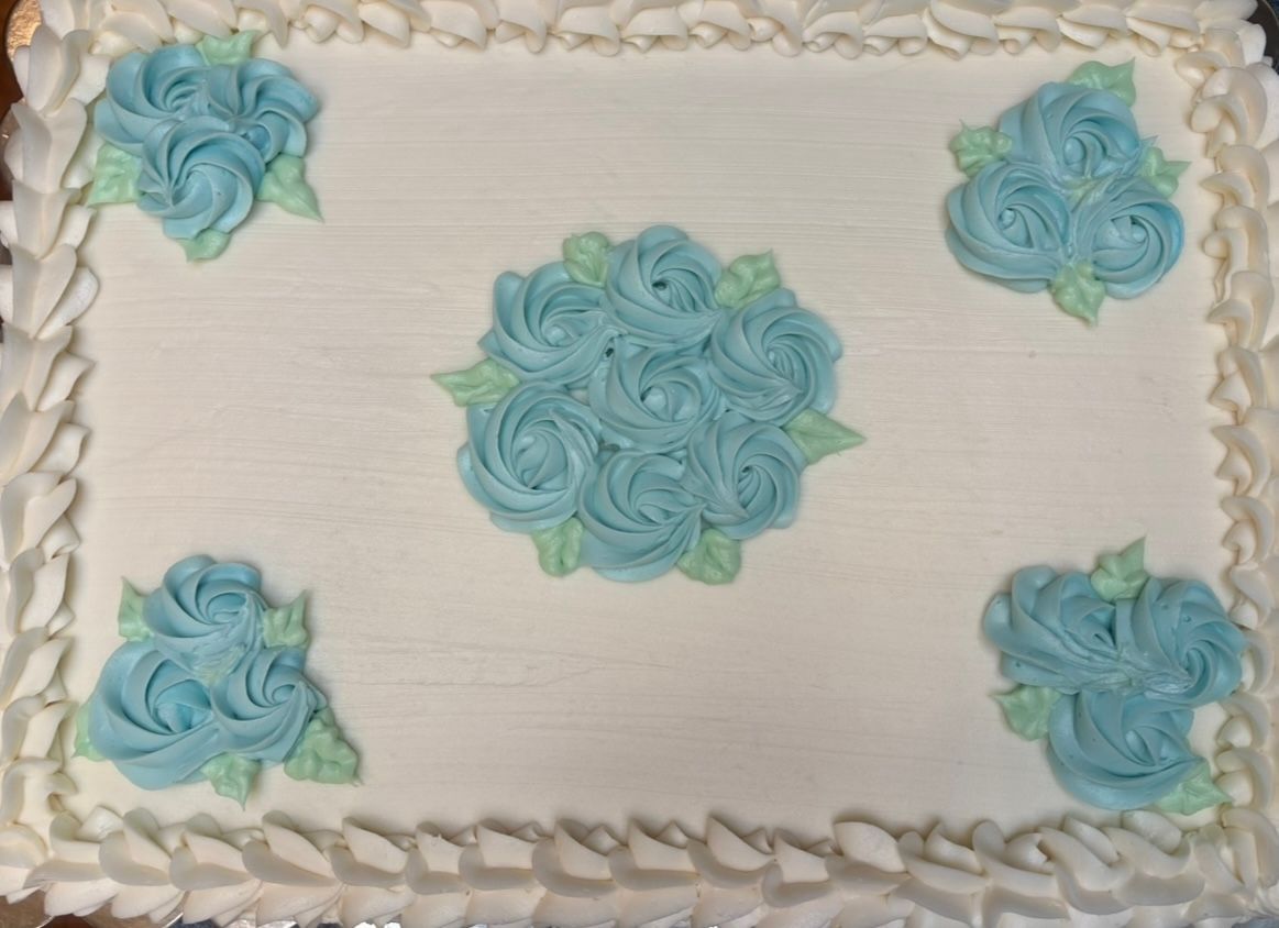 Rectangular cake with white frosting and blue frosting roses; white scalloped border. Base Camp Bakery Gunnison, Colorado https://share.google/AgqUzUKNzsb0ThlP5, https://www.facebook.com/BaseCampBakery1, https://www.instagram.com/basecampbakery_81230