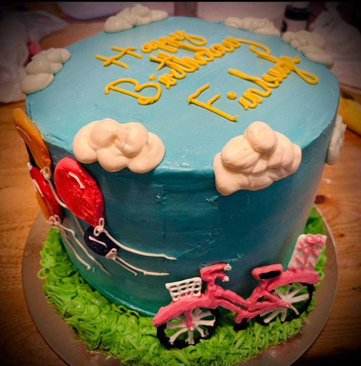 Birthday cake with blue frosting, clouds, balloons, and pink bicycle. Base Camp Bakery Gunnison, Colorado https://share.google/AgqUzUKNzsb0ThlP5, https://www.facebook.com/BaseCampBakery1, https://www.instagram.com/basecampbakery_81230