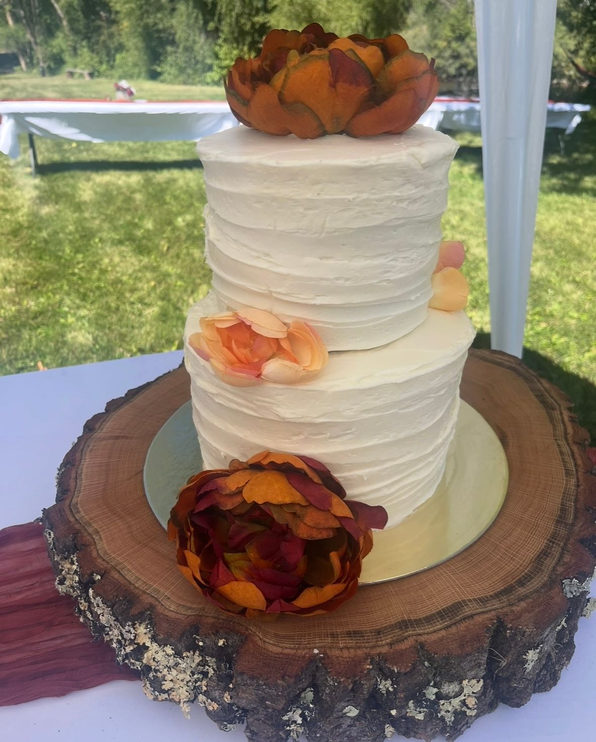 Two-tiered white frosted cake with fall-colored flowers on a wood slice. Outdoors, sunny setting. Base Camp Bakery Gunnison, Colorado https://share.google/AgqUzUKNzsb0ThlP5, https://www.facebook.com/BaseCampBakery1, https://www.instagram.com/basecampbakery_81230