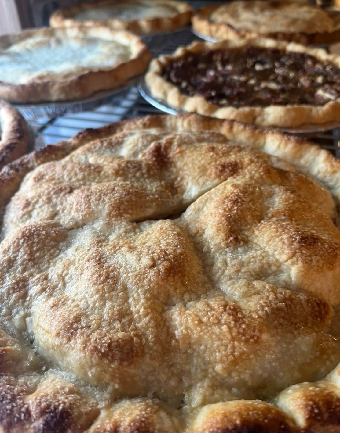 Close-up of a golden-brown pie crust with several other pies on a wire rack in the background. Base Camp Bakery Gunnison, Colorado https://share.google/AgqUzUKNzsb0ThlP5, https://www.facebook.com/BaseCampBakery1, https://www.instagram.com/basecampbakery_81230