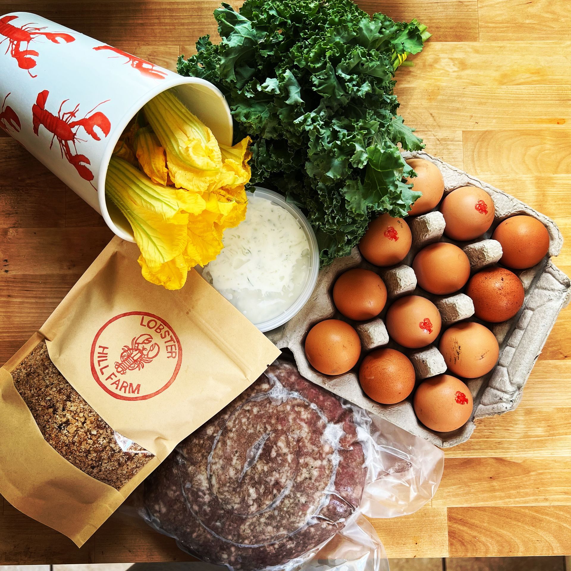 Fresh groceries: squash blossoms in a cup, kale, eggs, cheese, meat patty, and a bag of grain on a wooden surface.