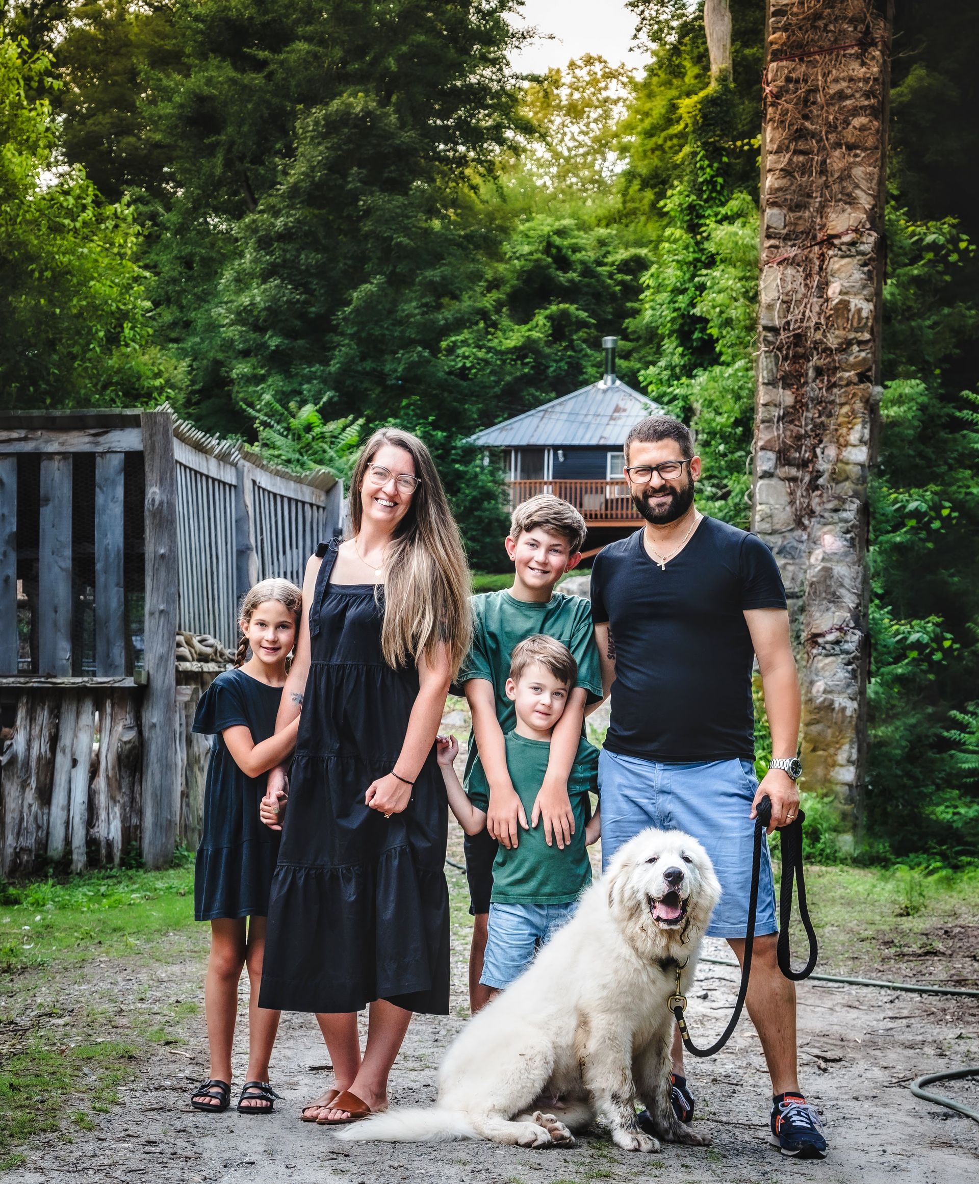 Family poses with a large dog outdoors; children, mother, and father smiling. Green trees and a small cabin in the background.