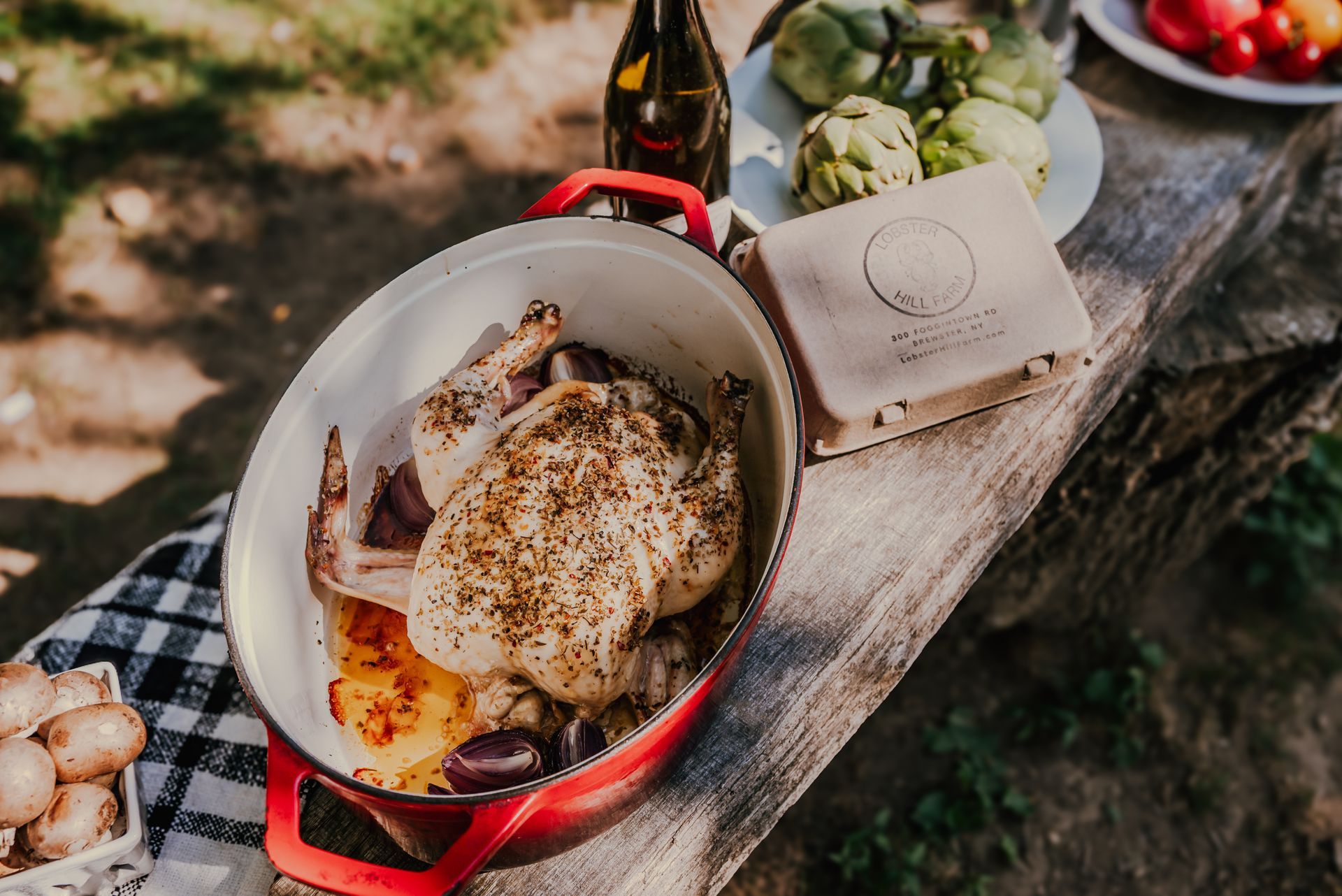 Roasted chicken in red pot outdoors, artichokes, vegetables, and beer on a wooden table.