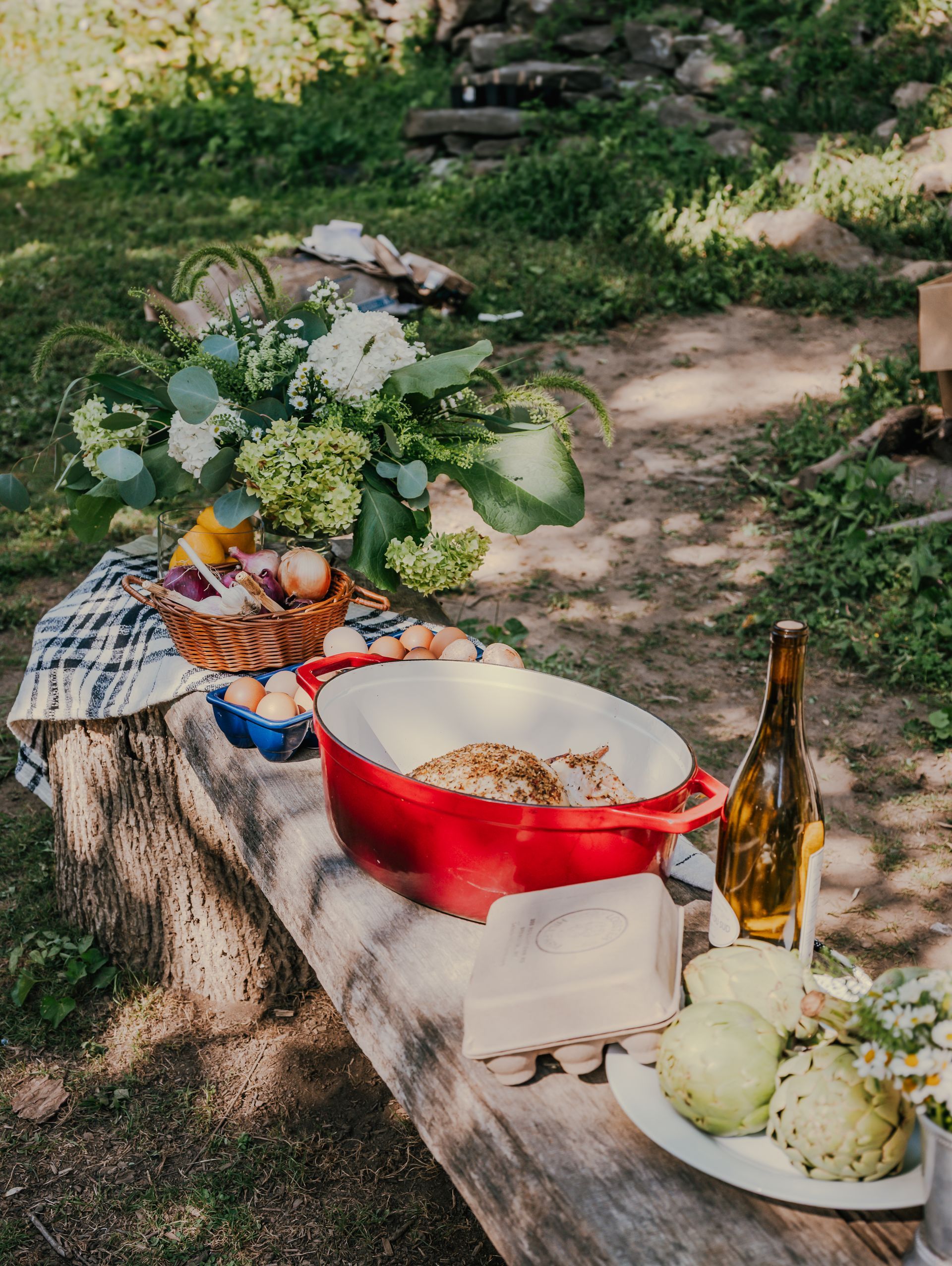 Outdoor picnic table with food and flowers. Red cast iron pot, eggs, wine, and fruit.