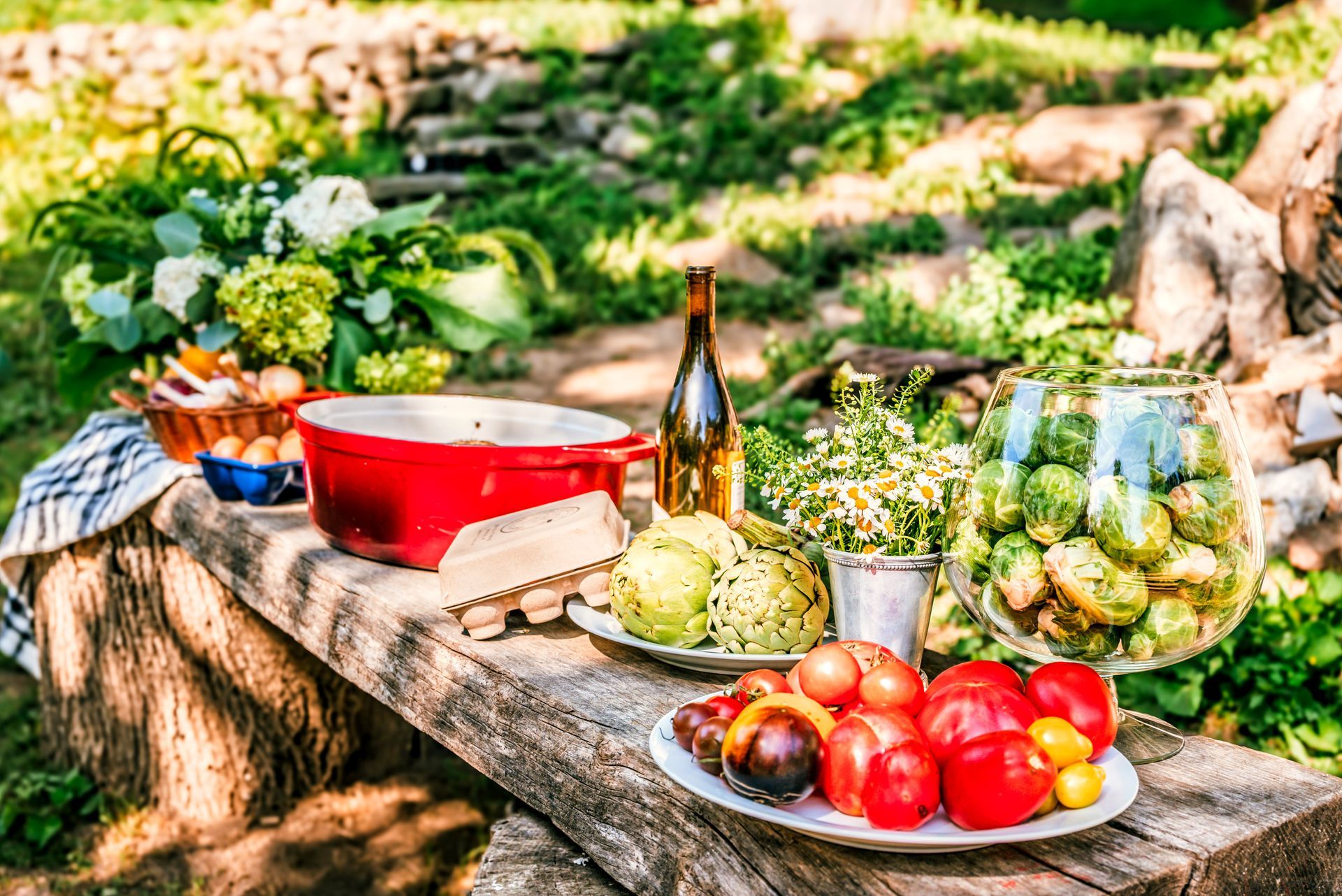 Outdoor table set for a meal with red pot, vegetables, and flowers in a garden setting.