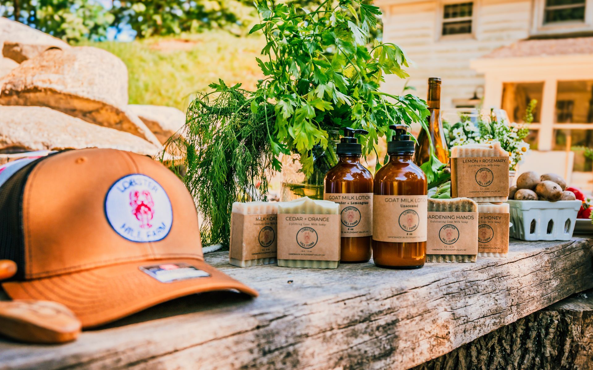 A brown cap, natural soaps, and bottles sit on a wooden table, with greenery and a house in the background.