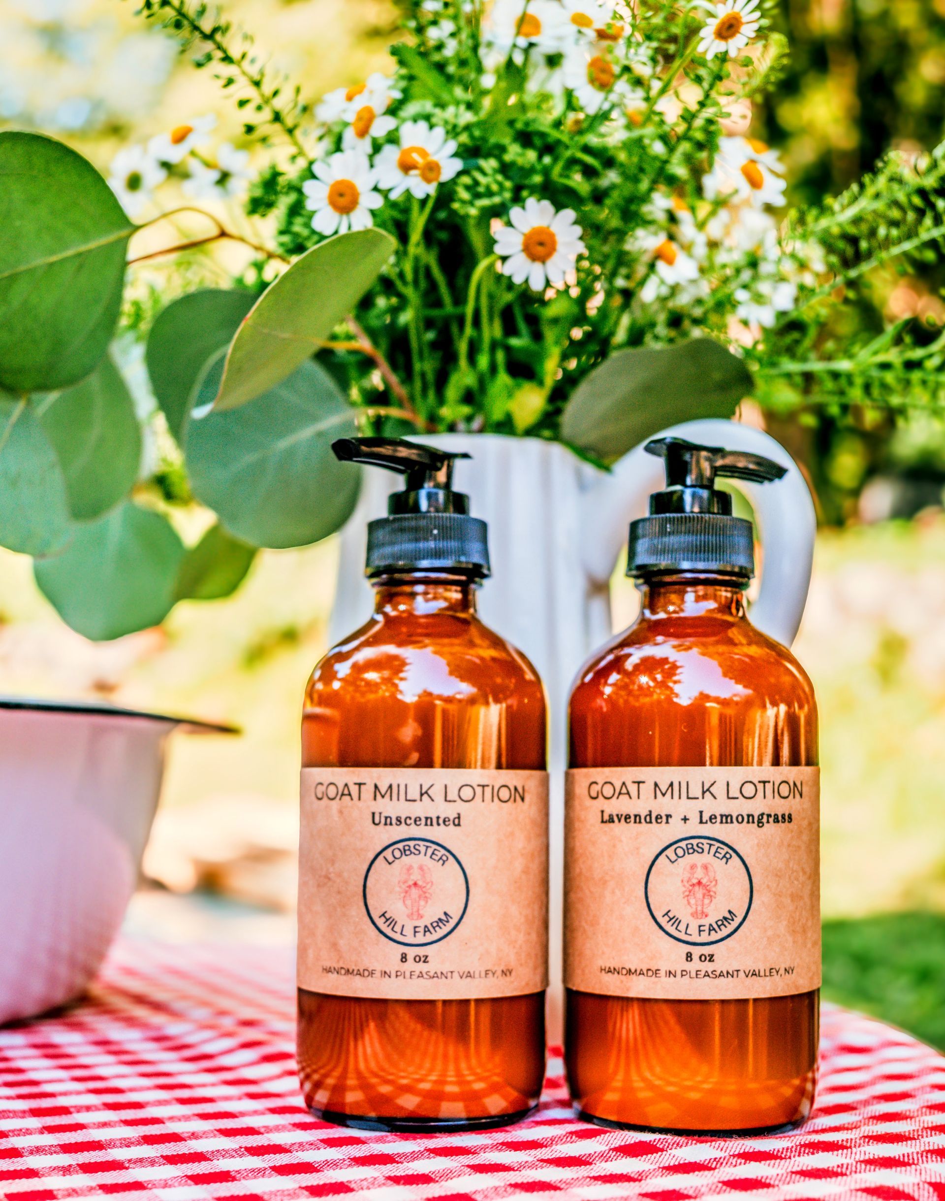 Two amber-colored goat milk lotion bottles with paper labels on a red-checkered table, flowers in the background.