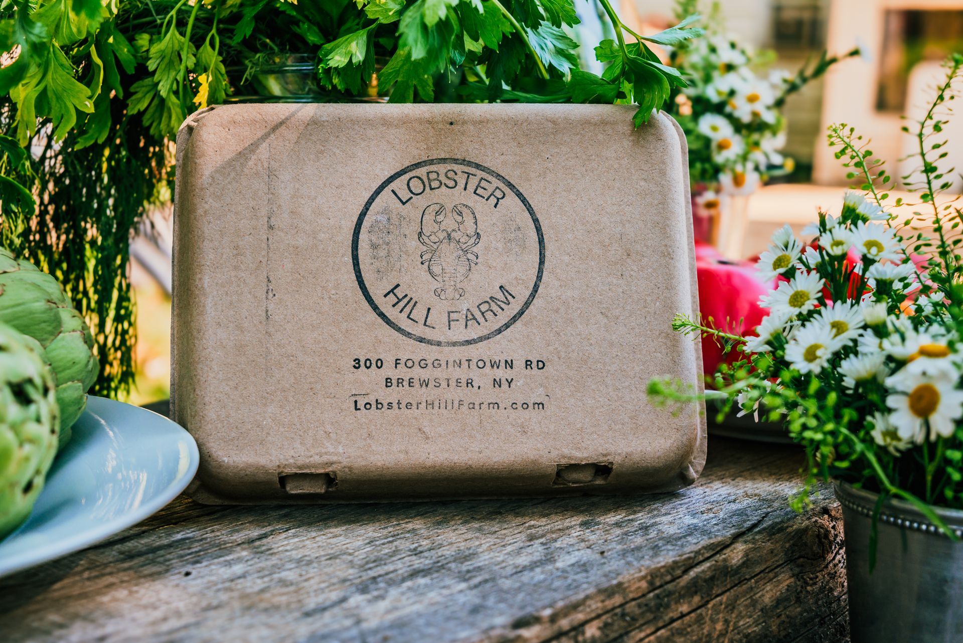 Brown box with Lobster Hill Farm logo on a weathered wooden surface, with greenery and flowers.