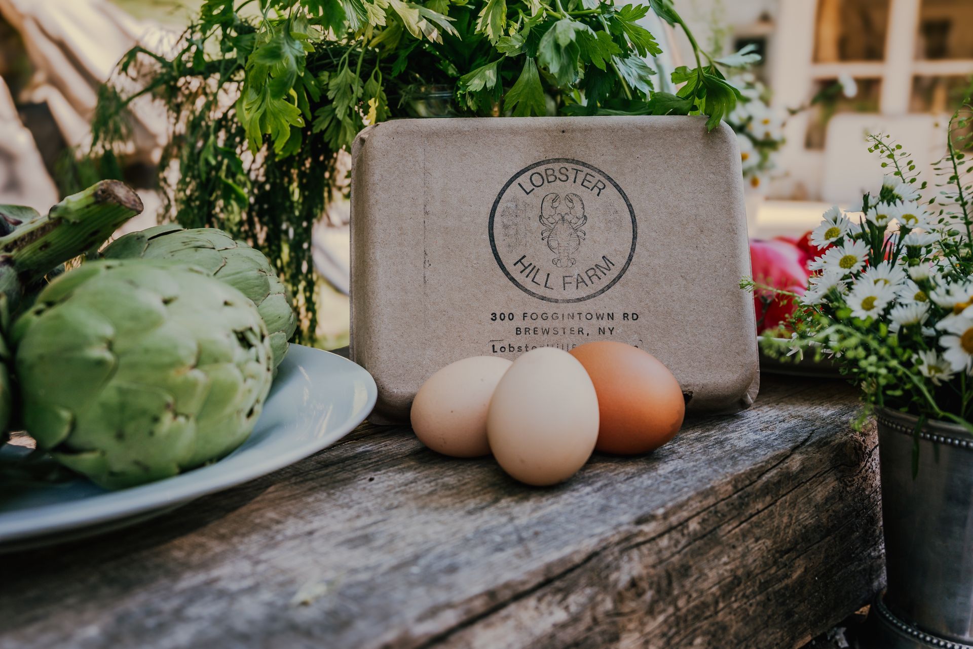 Eggs, artichokes, and egg carton on a wooden surface with greenery in the background.