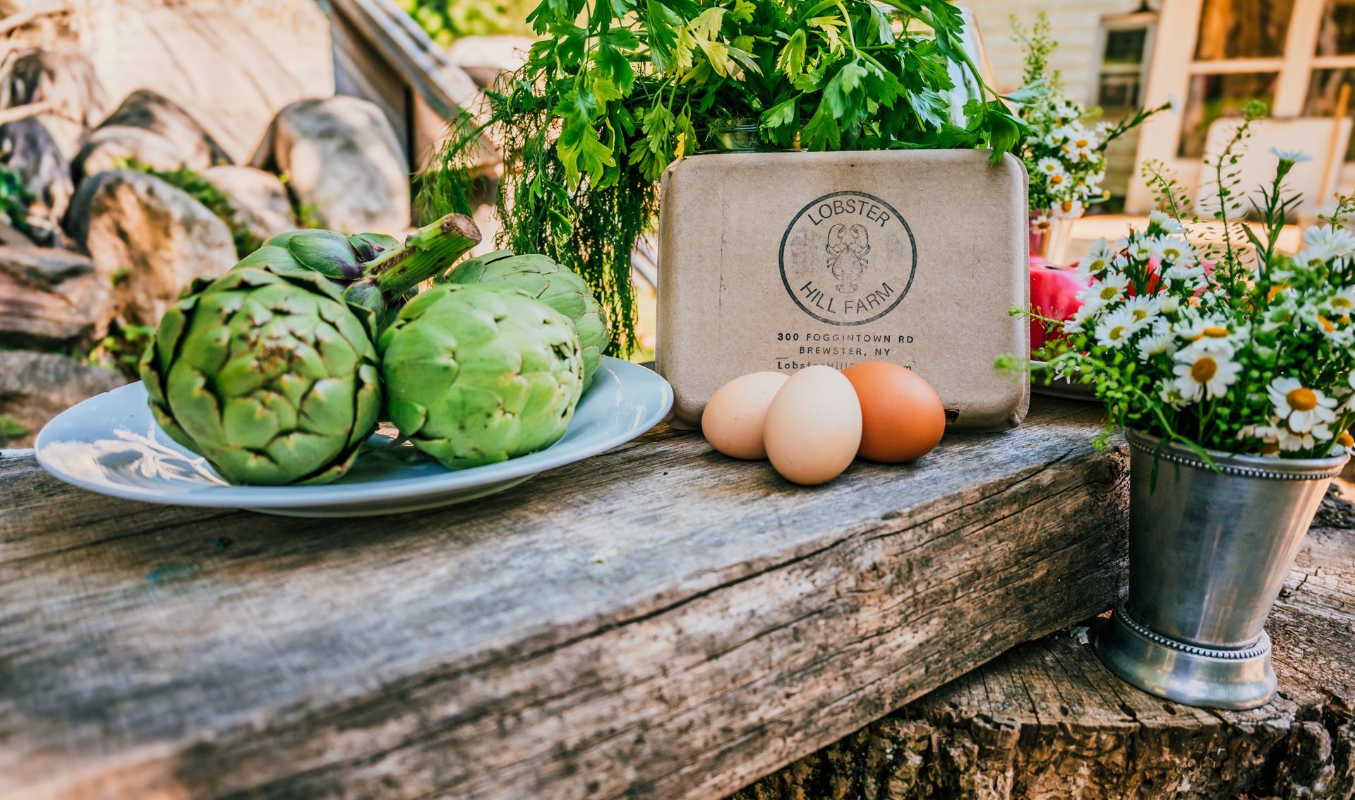 Artichokes, eggs, and flowers on a rustic wooden surface with a backdrop of greenery and a building.