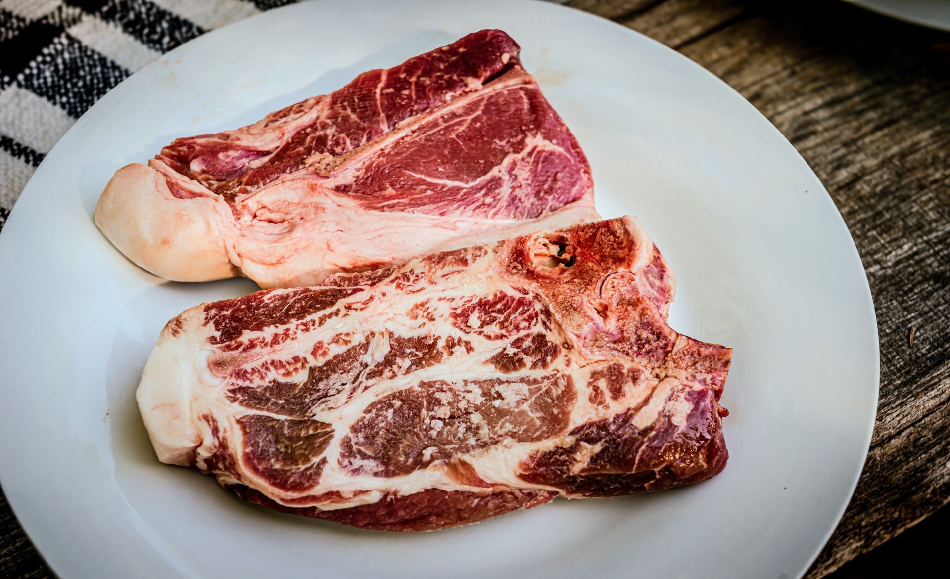 Two raw pork steaks on a white plate, set on a wooden surface with a checkered napkin.