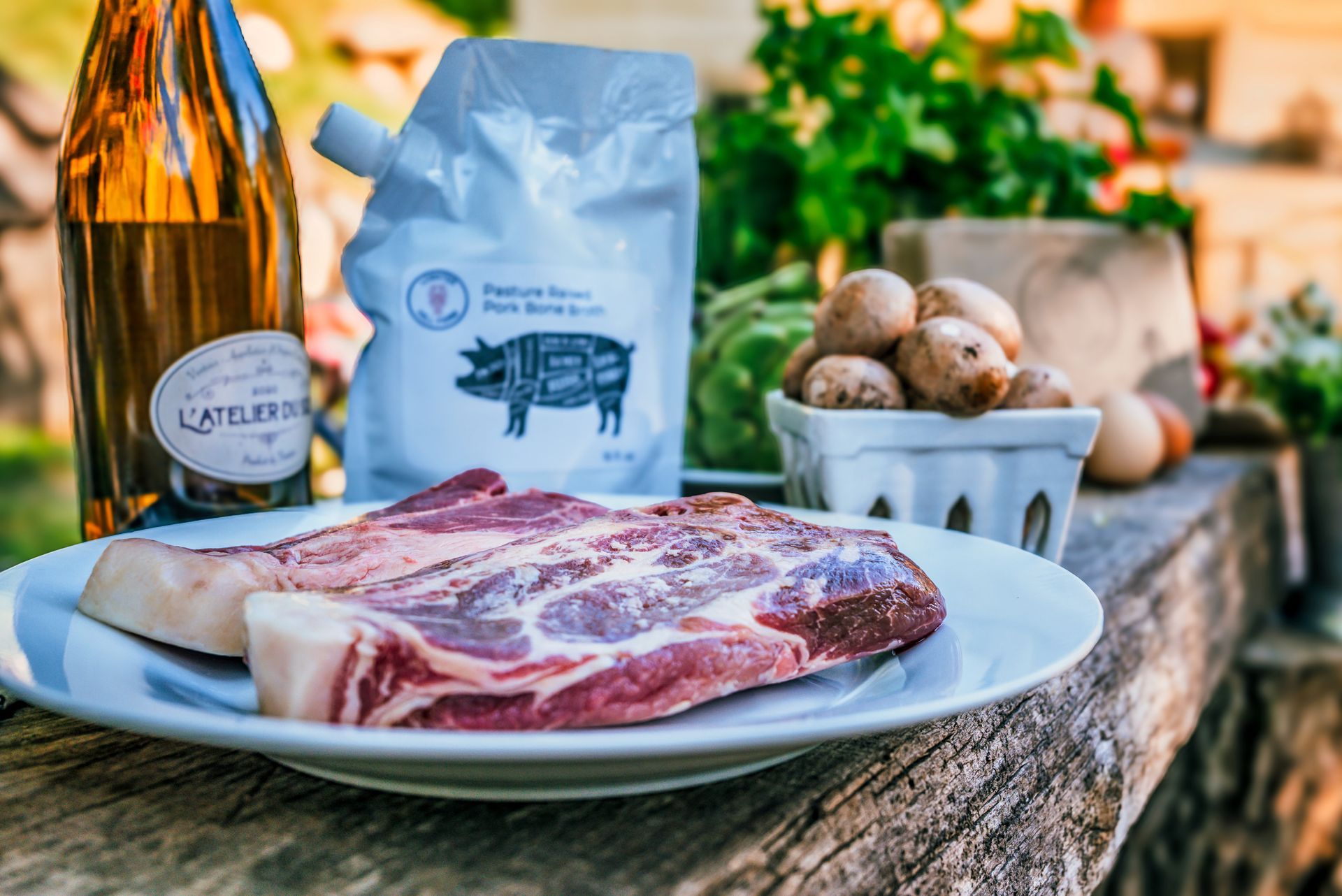 Close-up of a plate with raw pork chop, ingredients for grilling outdoors: bottle of wine, salt pouch, mushrooms, and herbs.