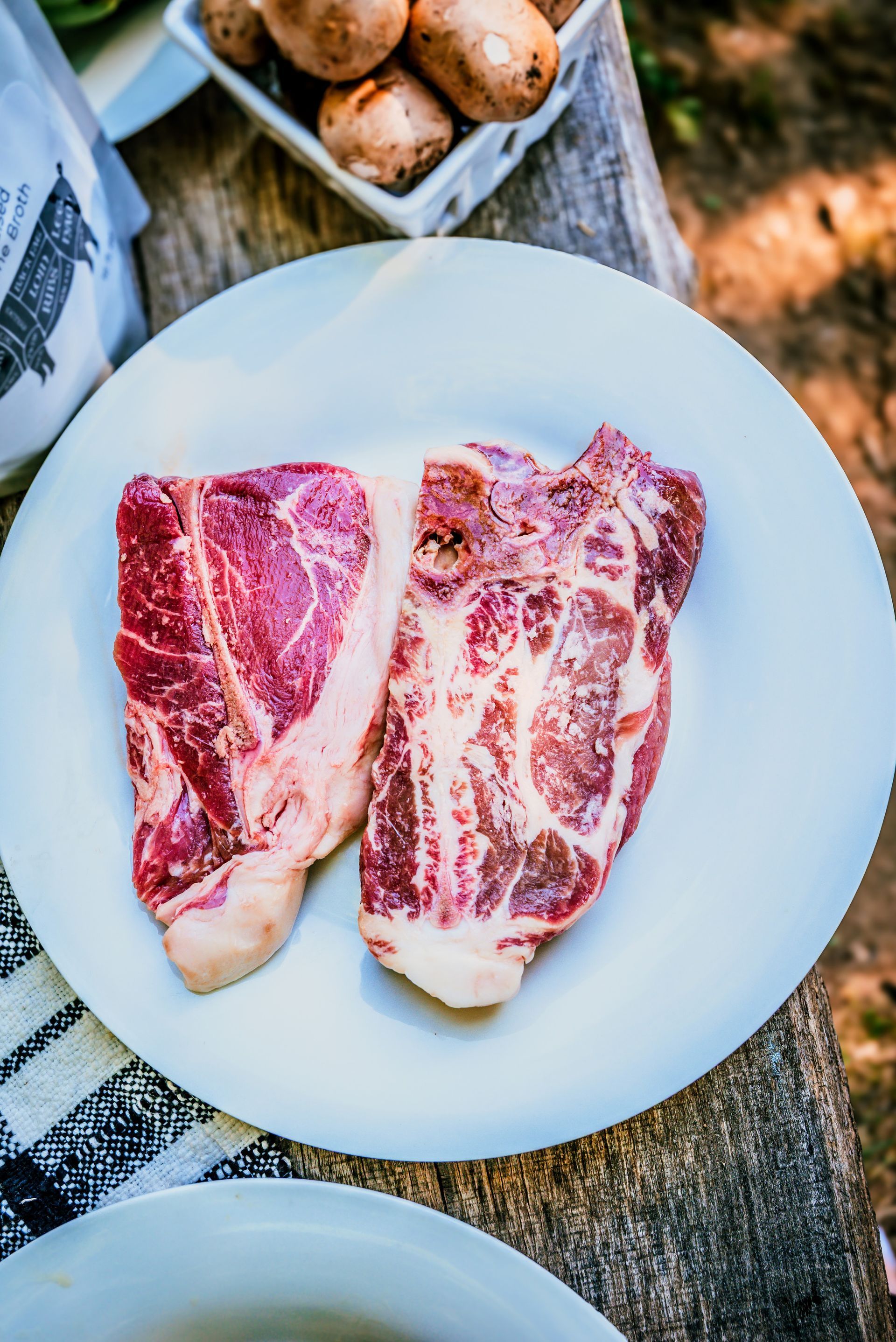 Two raw, marbled steaks on a white plate, with mushrooms in a bowl nearby, outdoor setting.