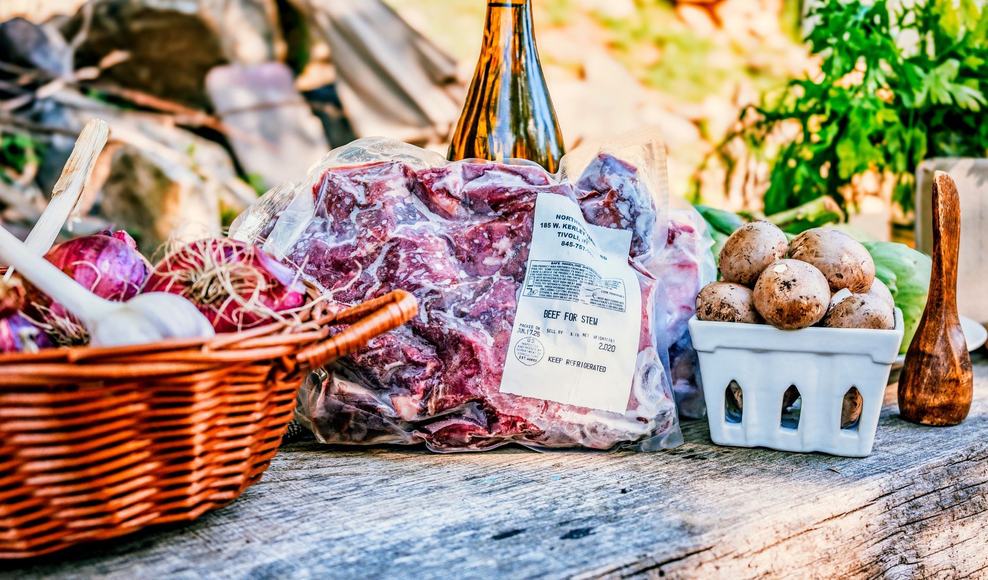 Basket of vegetables, vacuum-sealed meat, mushrooms, and bottle on rustic table outdoors.