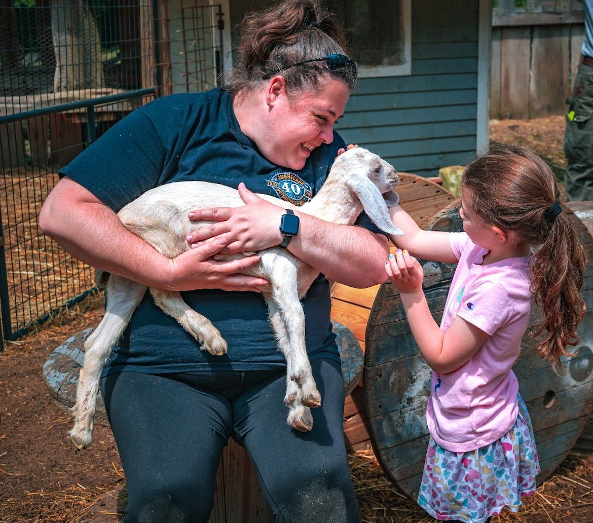 Woman holding a white baby goat, smiling at a young girl reaching to pet it outdoors.