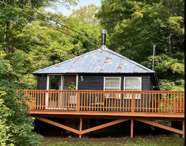 Black cabin with a weathered metal roof, a wooden deck, and surrounded by trees.